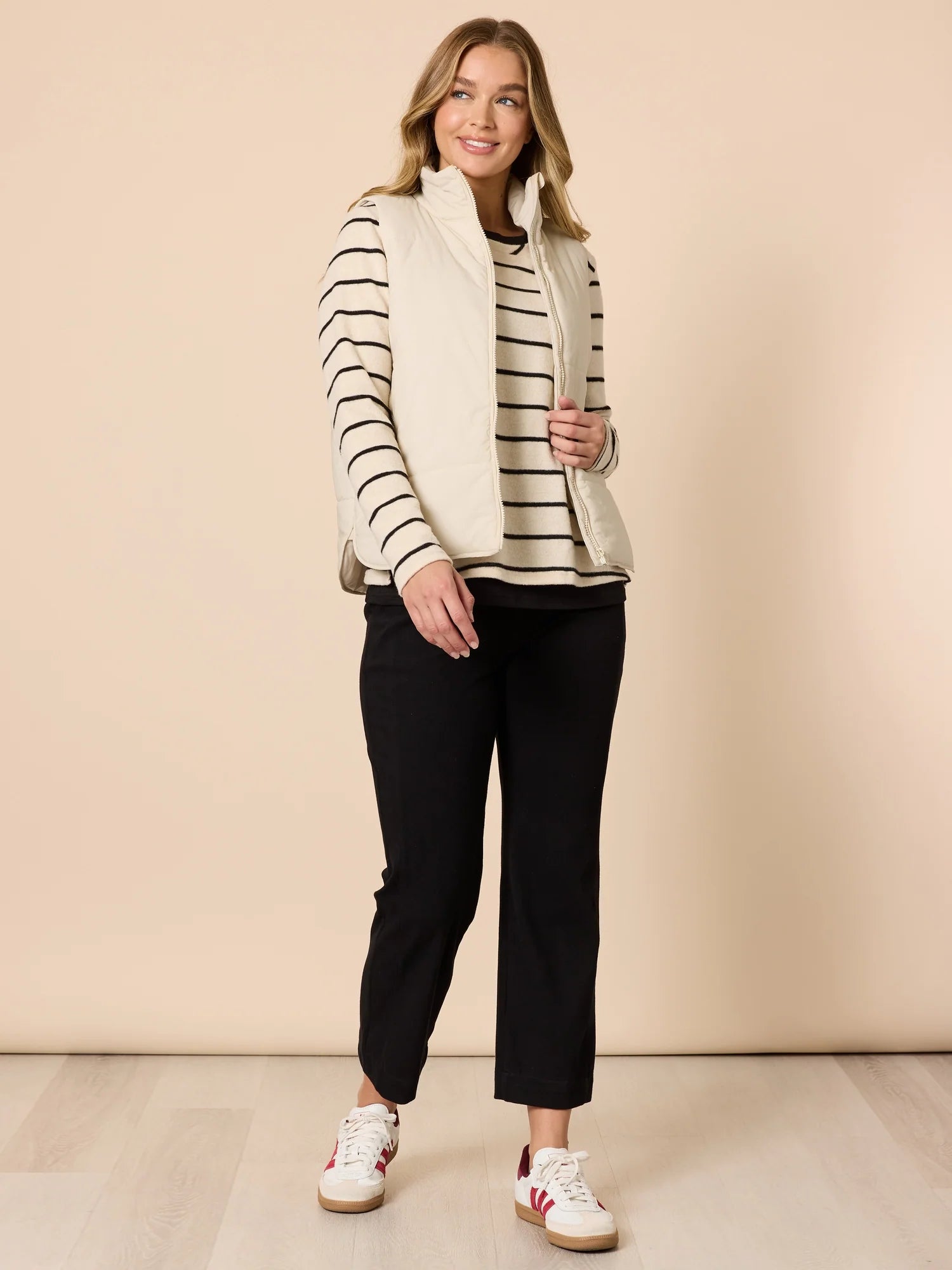 Image of Woman wearing a Threadz striped sweater, black pants, and white sneakers against a beige background