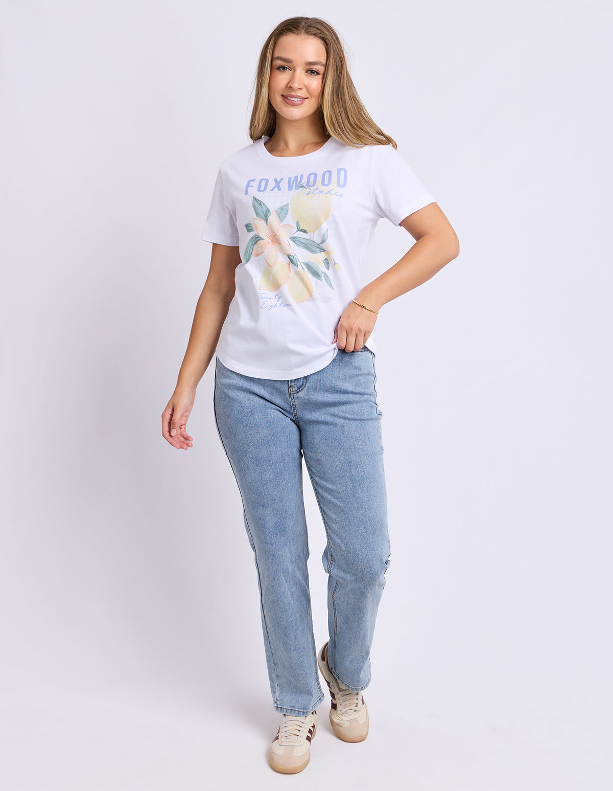 Woman wearing a white t-shirt with a graphic design with flowers and lemons and blue jeans on a plain background