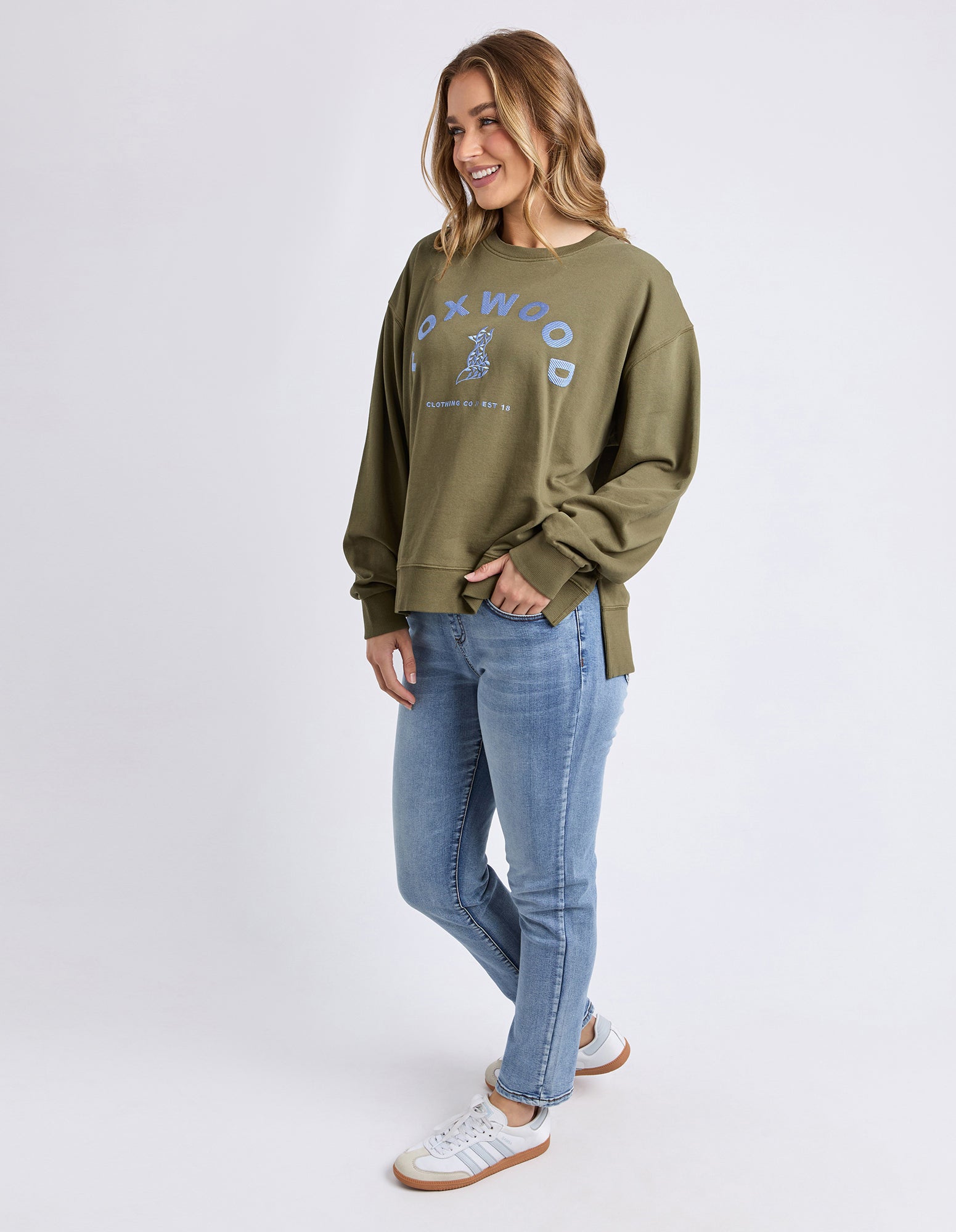 Woman wearing a green jumper with branding and blue jeans on a white background