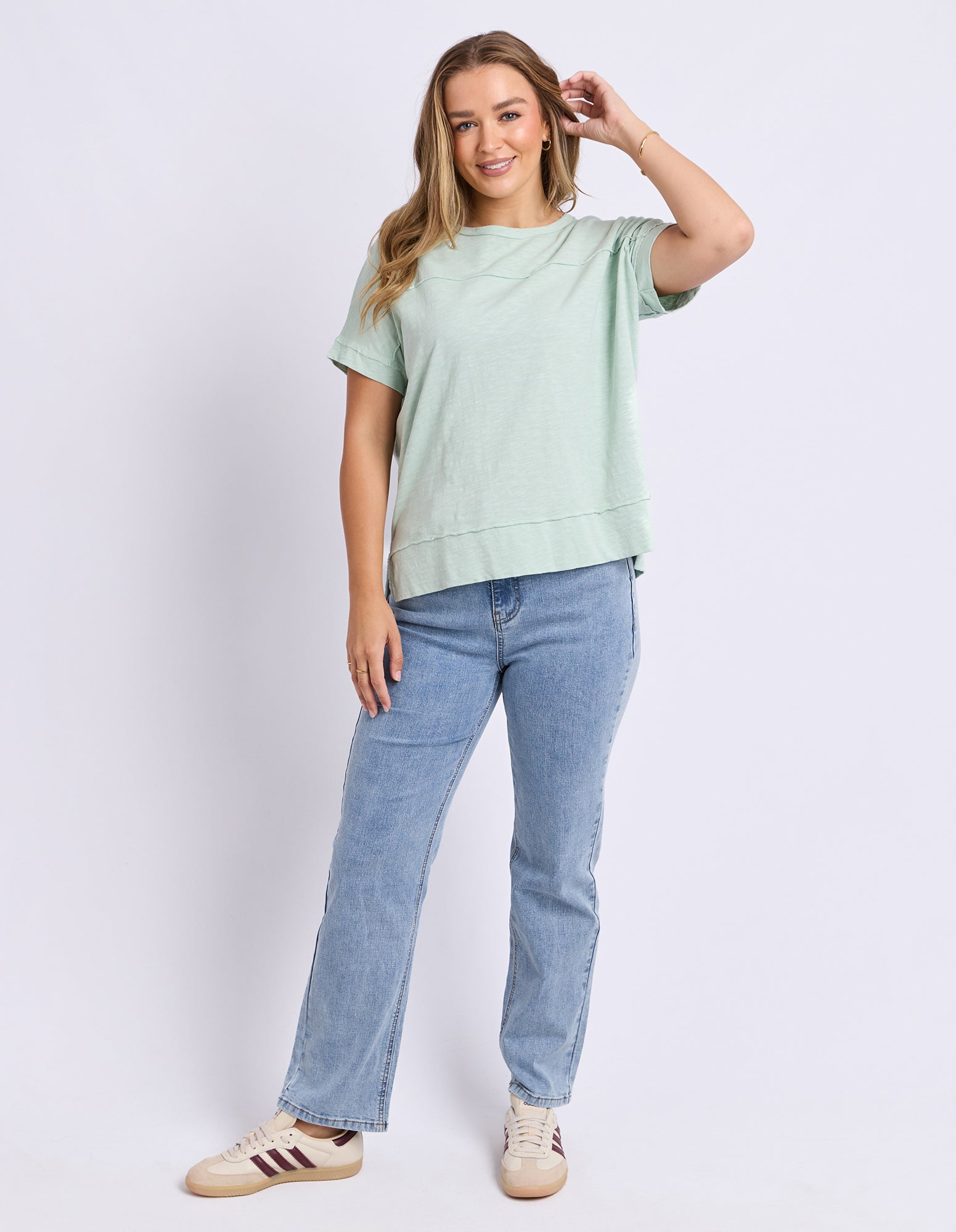 A woman wearing a light sage green short-sleeve round neck tee and light-wash blue jeans, paired with beige sneakers, against a plain light grey studio background.