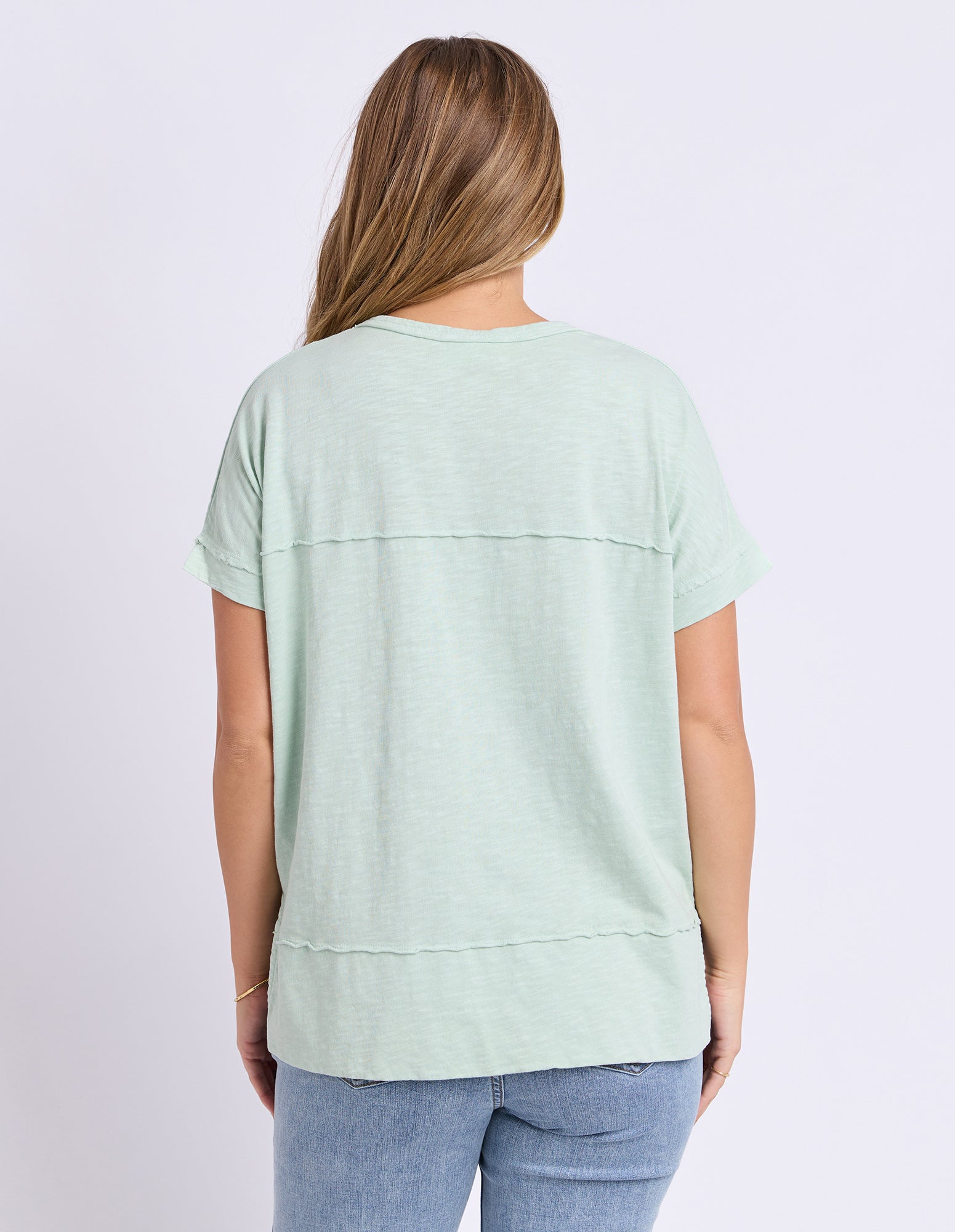 A woman viewed from the back wearing a light green short-sleeve top and blue jeans against a plain light background.