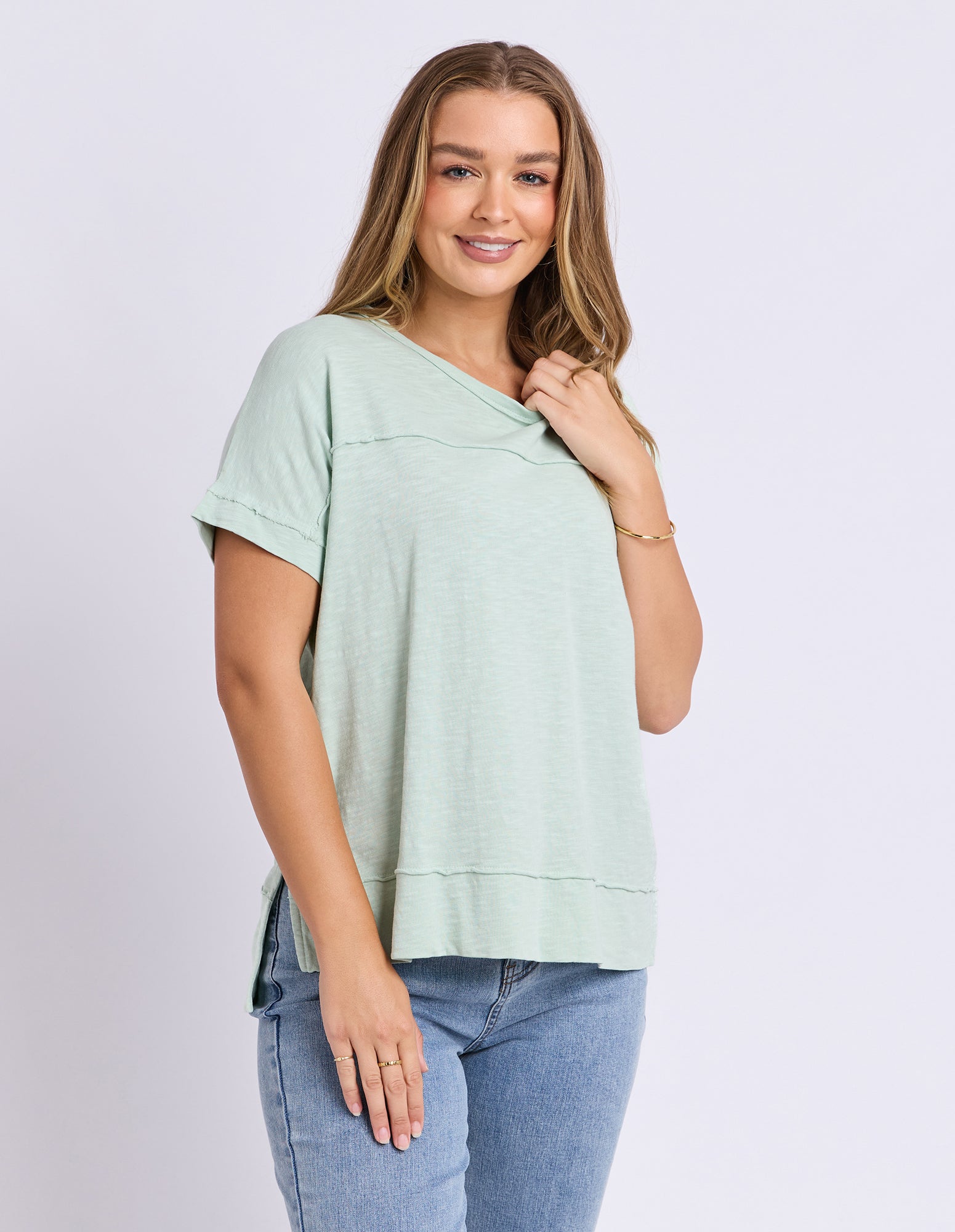 Image of A woman modeling a light sage green short-sleeve tee with a relaxed fit, paired with blue jeans, against a plain light grey studio background.