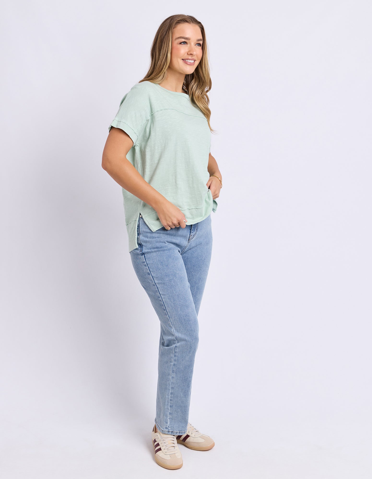 Image of A woman wearing a light green short-sleeve t-shirt and light blue jeans, standing against a plain light grey studio background.