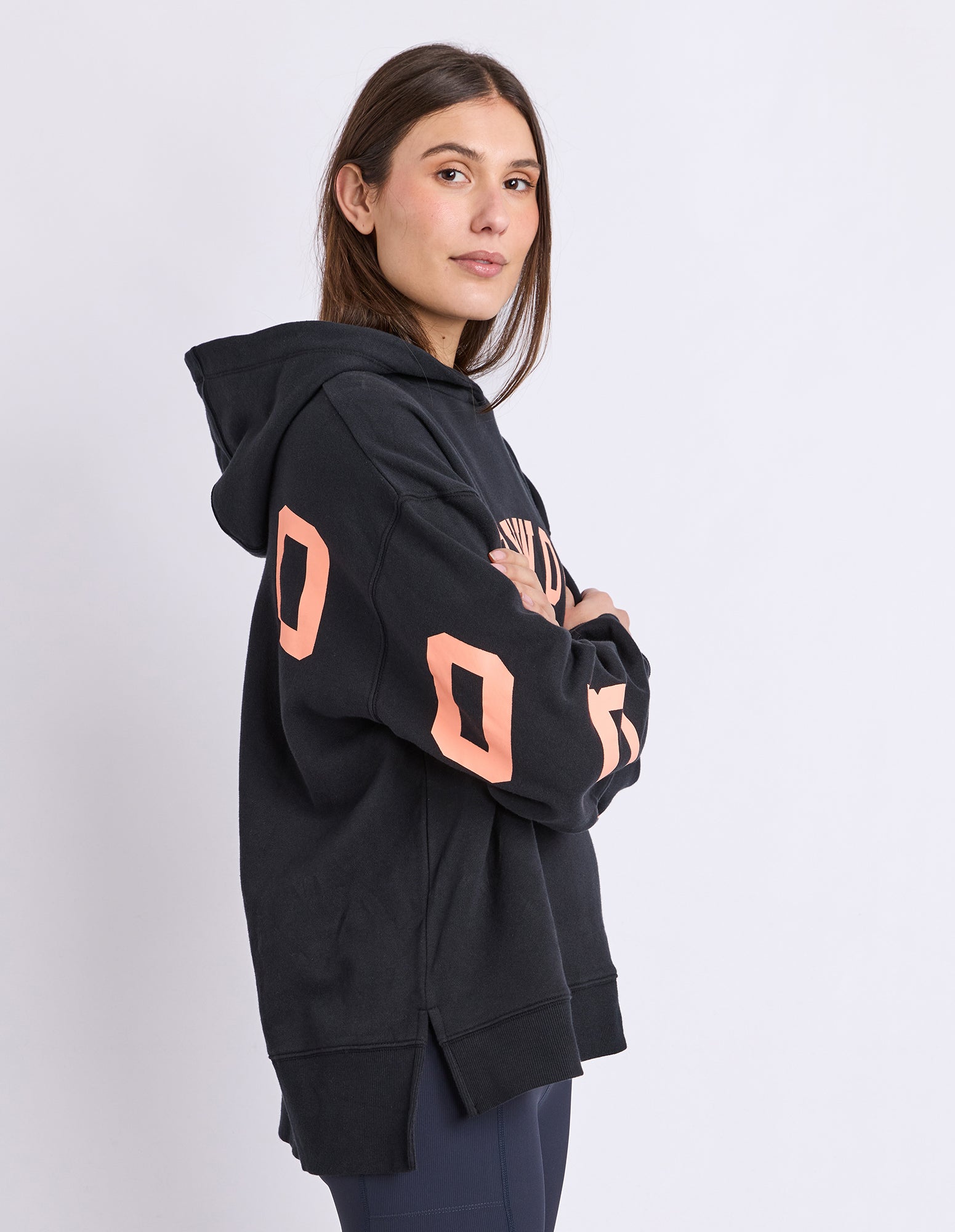 A woman modelling a black oversized hoodie with large orange coloured letters on the sleeves, photographed against a light grey studio background.