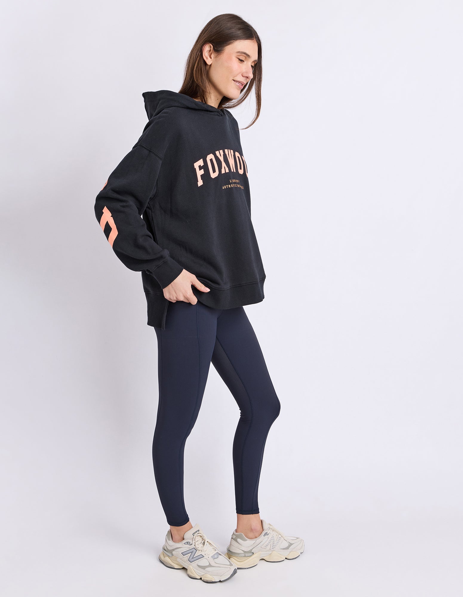 Image of A woman modelling a black oversized hoodie with 'FOXWOOD' printed in orange across the chest, paired with dark leggings and beige sneakers, against a light grey studio background.