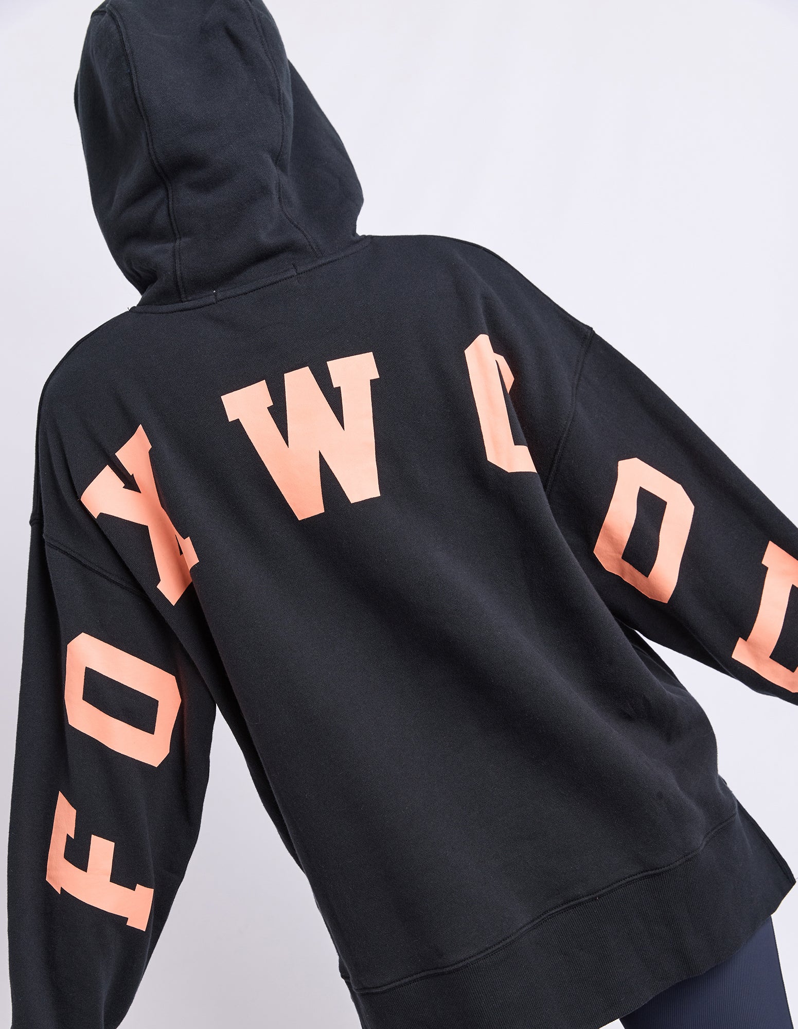 Image of A black hoodie shown from the back, featuring large orange coloured letters spelling 'FOXWOOD' across the upper back and sleeves, worn by a model against a light grey studio background.