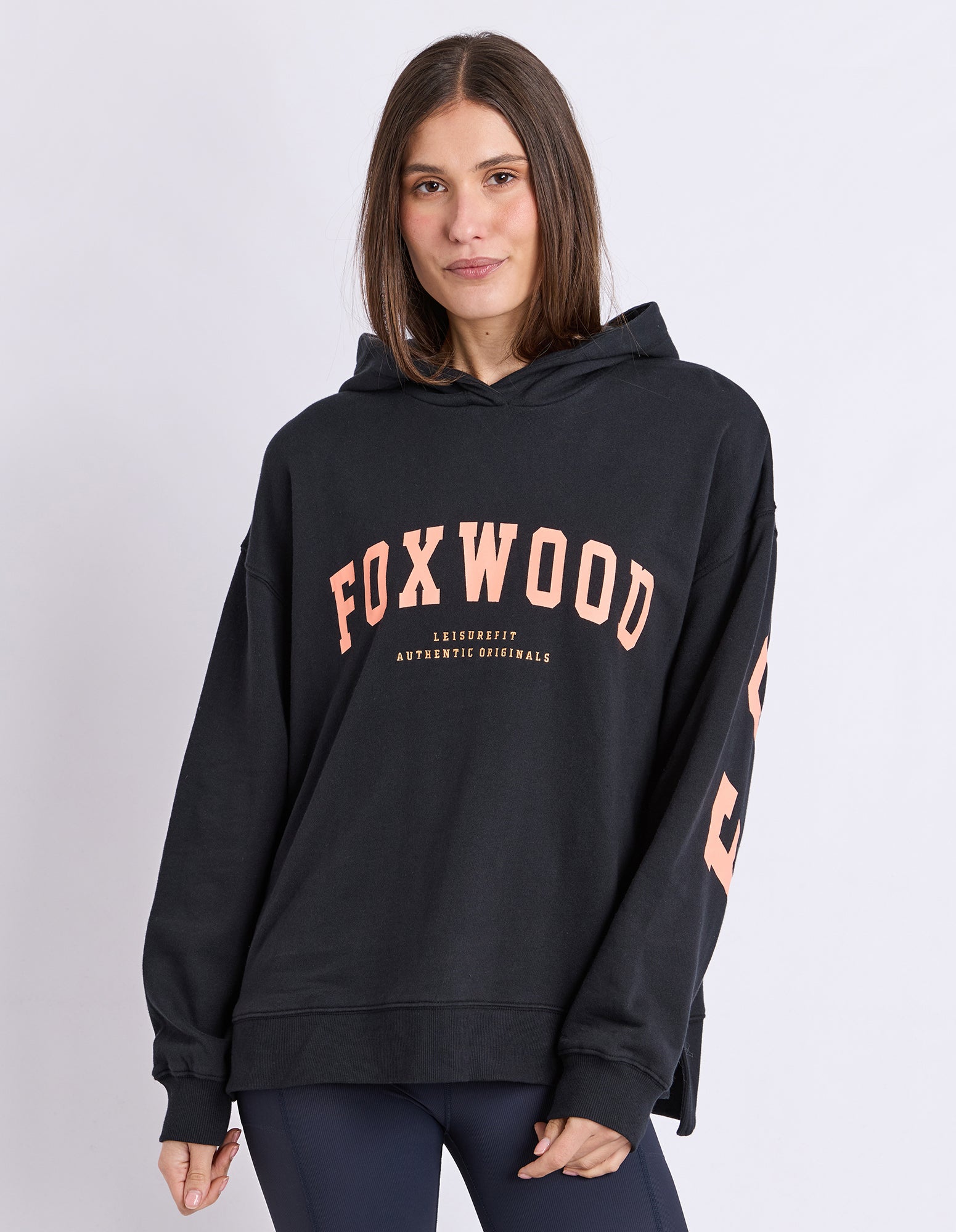 A woman wearing a black hoodie with 'FOXWOOD' printed in orange across the chest and matching lettering down the sleeve, posed against a light grey studio background.