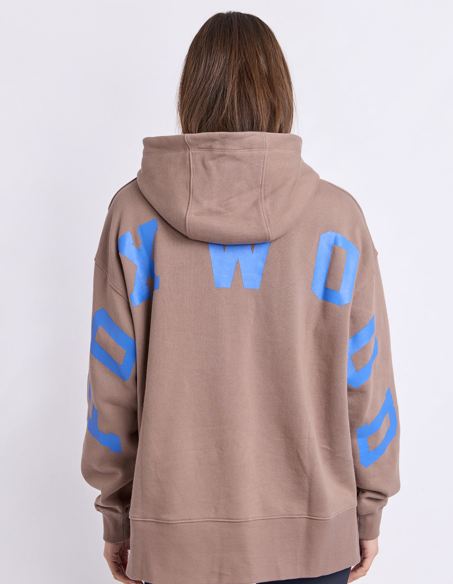 A woman shown from the back wearing an oversized brown hoodie with large blue block letters spelling 'FOXWOOD' across the back and down the sleeves, against a plain light grey studio background.