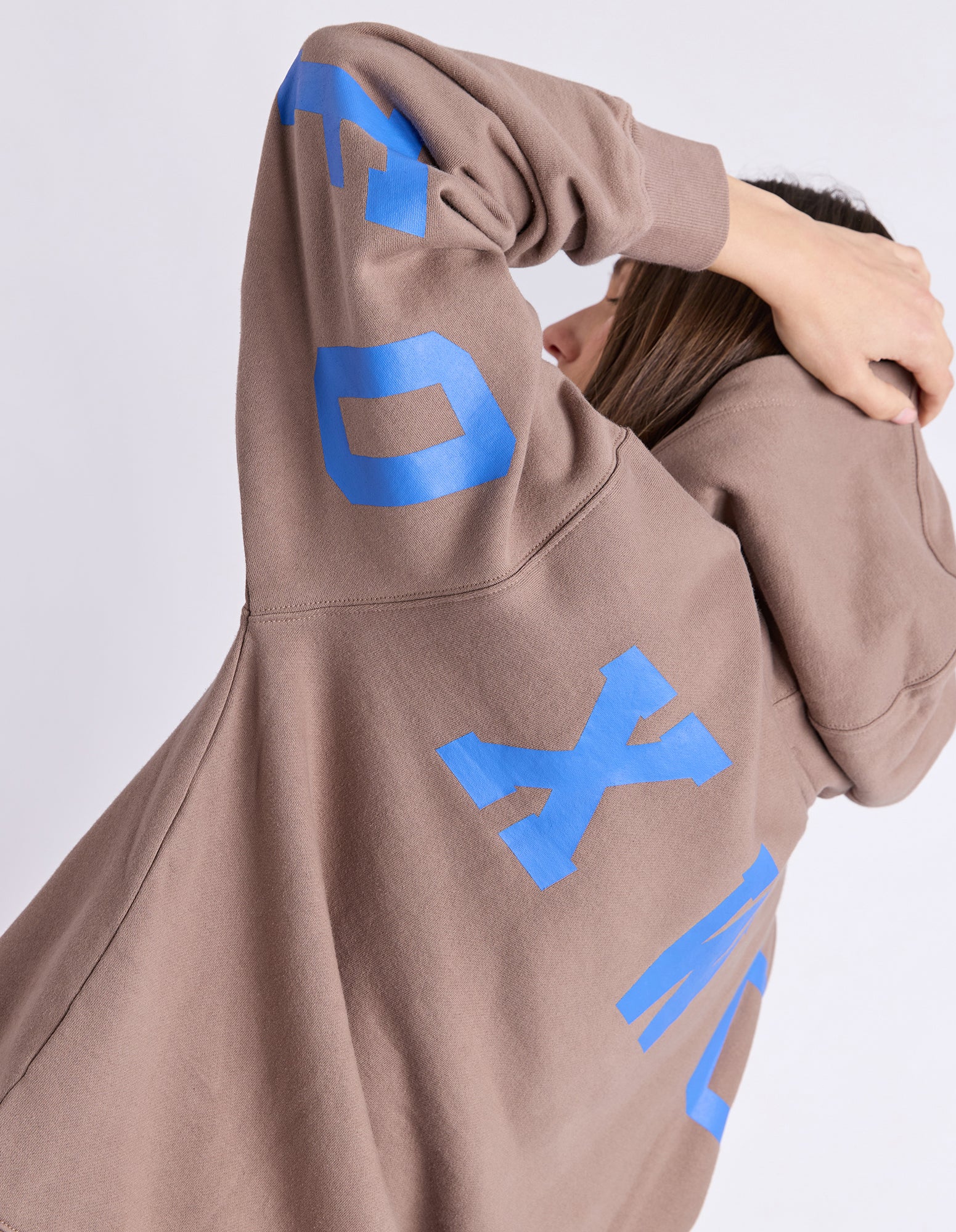 Image of A woman wearing an oversized brown hoodie with large blue graphic lettering on the back, shown from a side-back angle against a light grey studio background.