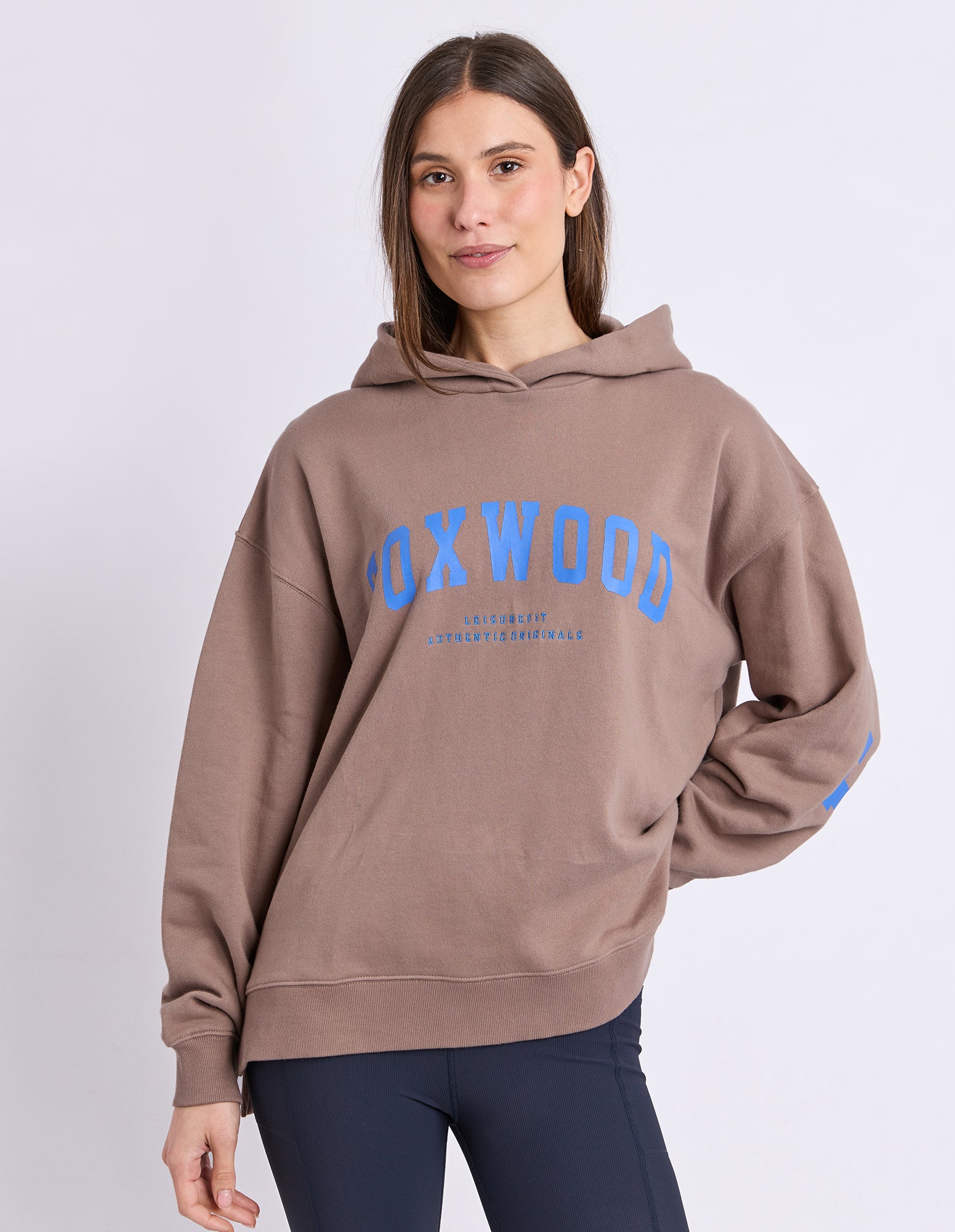 Image of A woman wearing a brown hoodie with a blue 'FOXWOOD' graphic on the chest, paired with dark navy leggings, against a light grey studio background.