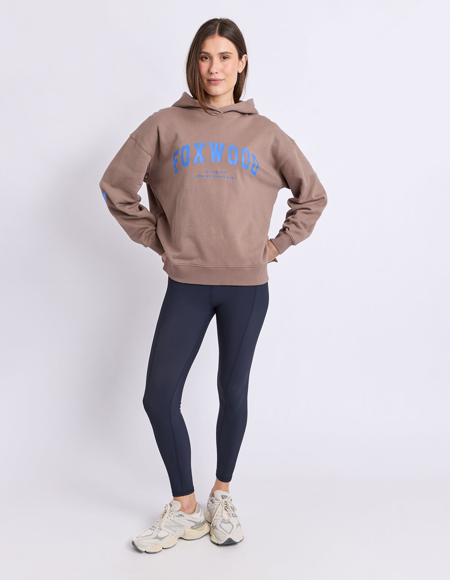 Image of A woman modeling a brown hoodie with a blue 'FOXWOOD' graphic on the chest, paired with dark navy leggings and beige sneakers, against a light grey studio background.