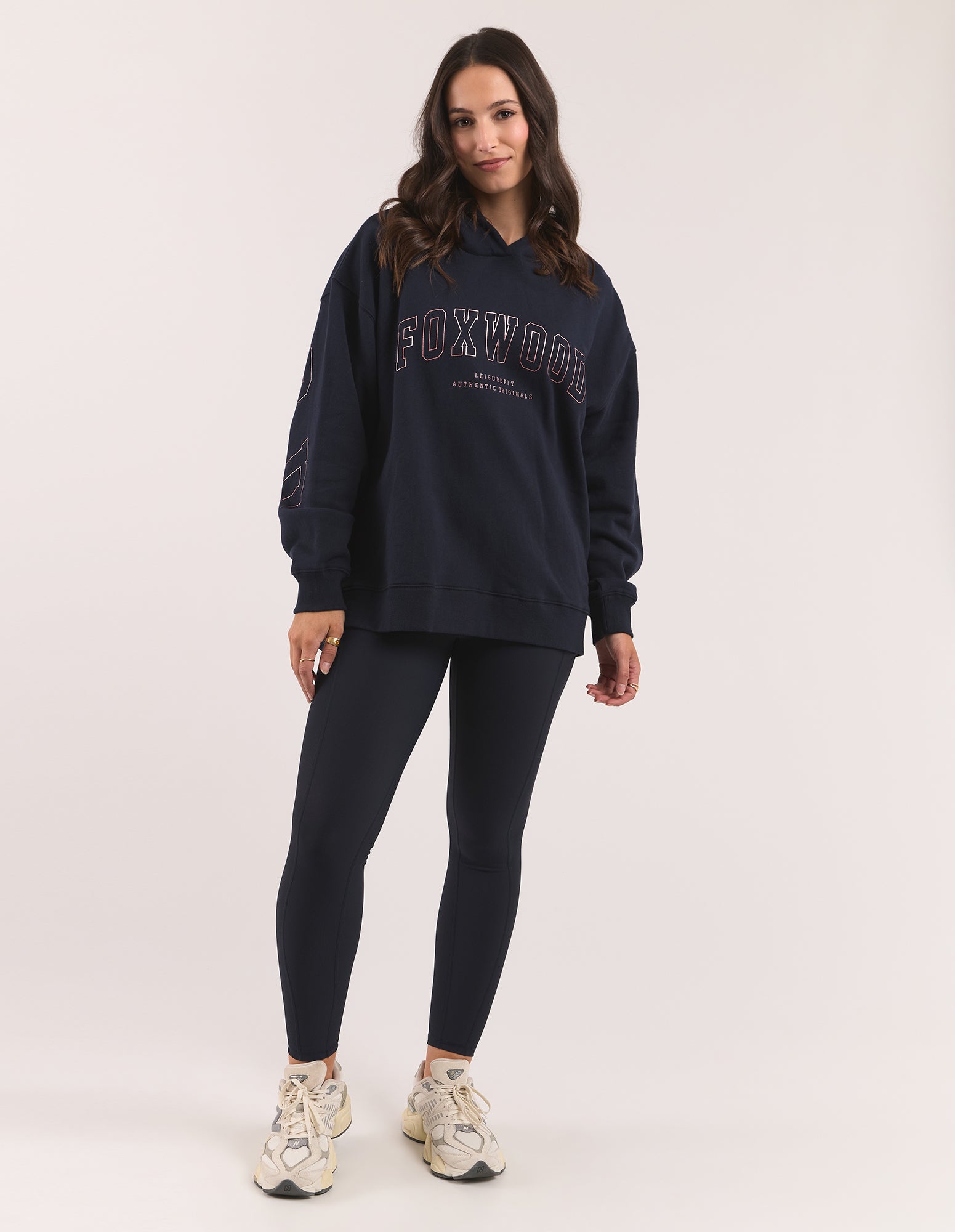 Image of A woman modeling a dark navy blue Foxwood hoodie wth leggings, paired with beige sneakers, against a plain light grey studio background.