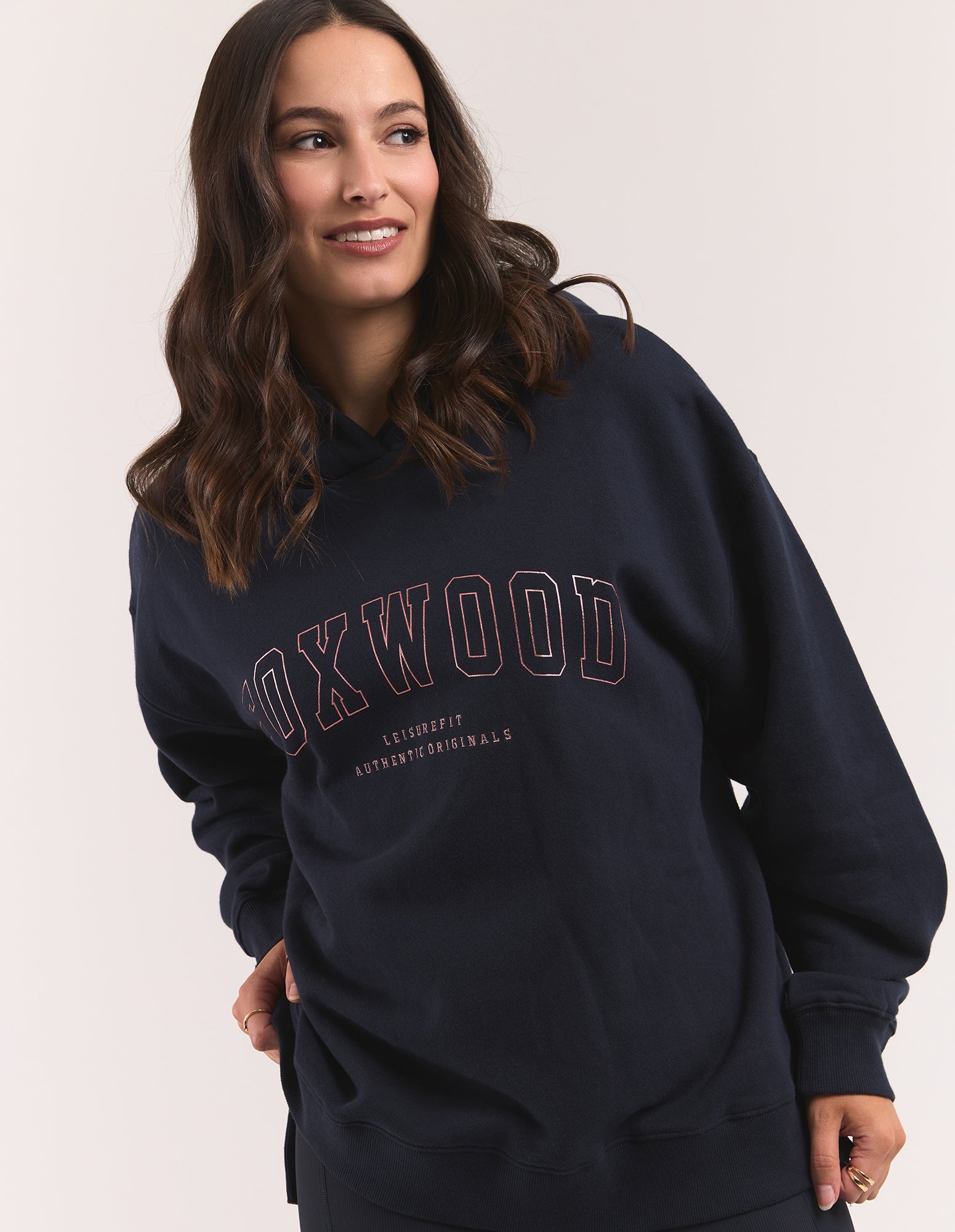 Image of A woman wearing a navy blue hooded jumper with 'FOXWOOD' printed in a thin pink outline across the chest, set against a plain light beige background.