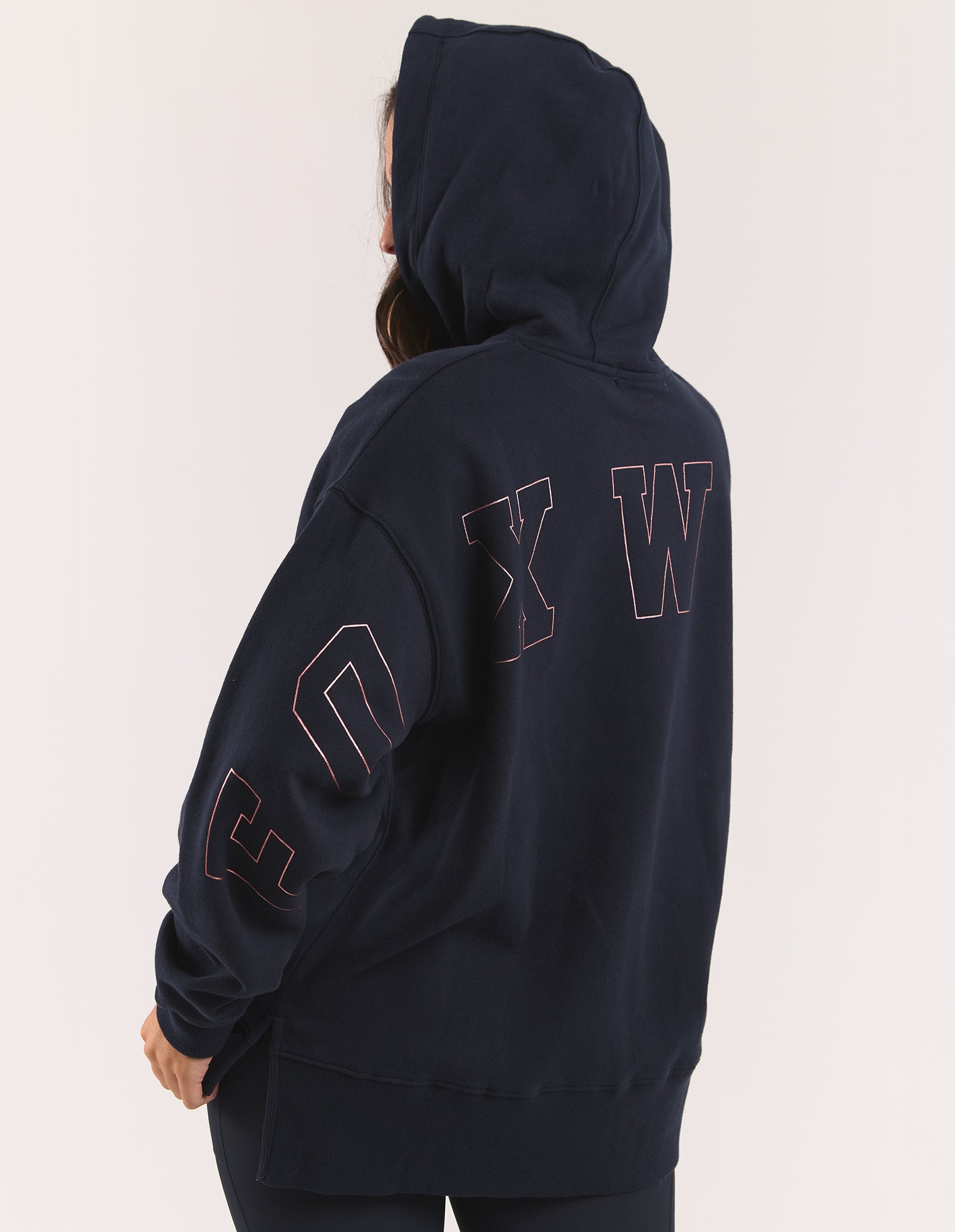 Image of A person shown from the back wearing a dark navy hooded jumper with large outlined lettering across the upper back and down the sleeves, photographed against a plain light background.