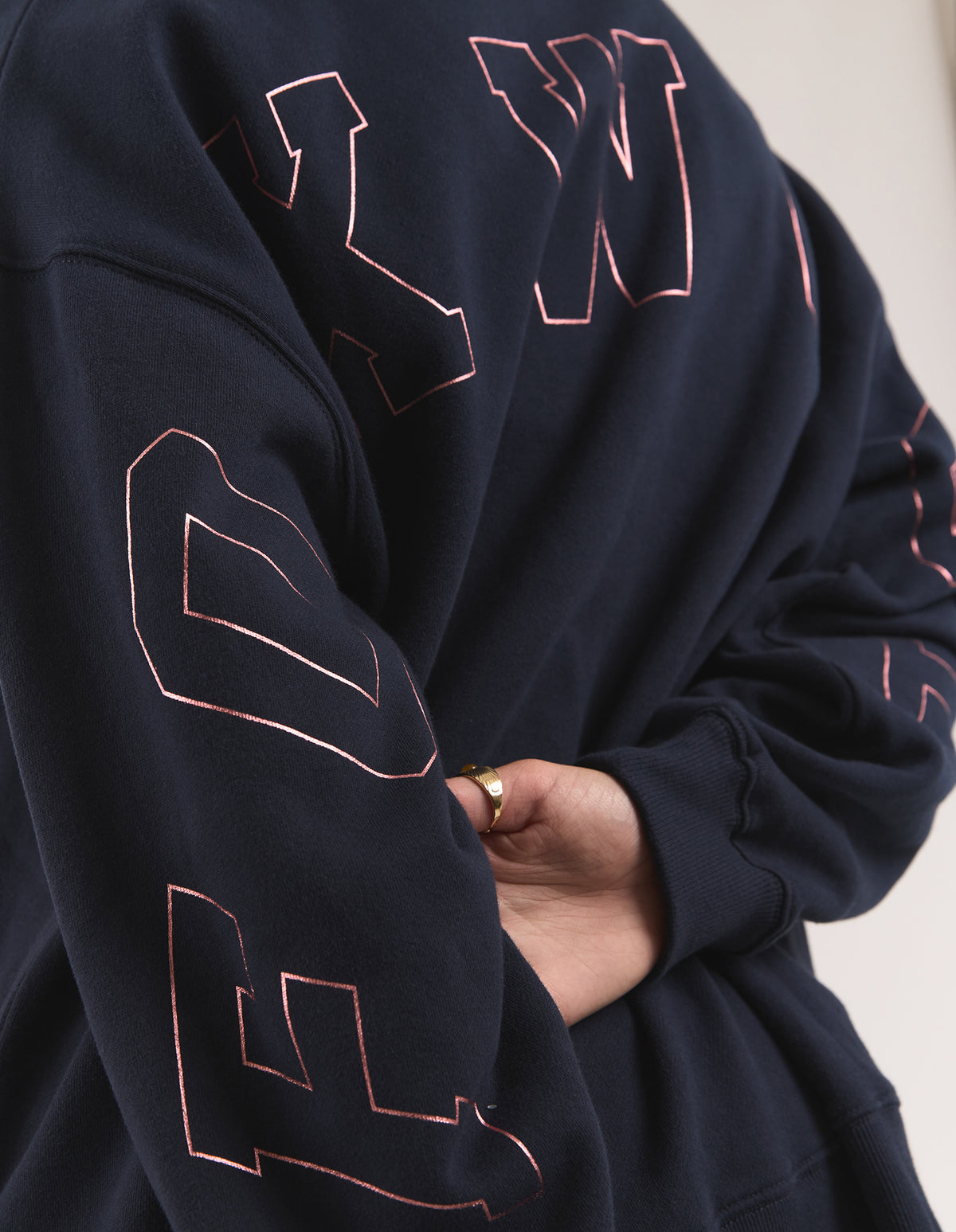 A close-up of a woman wearing a dark navy hoodie with large, rose gold outlined varsity-style letters across the front and sleeves. 