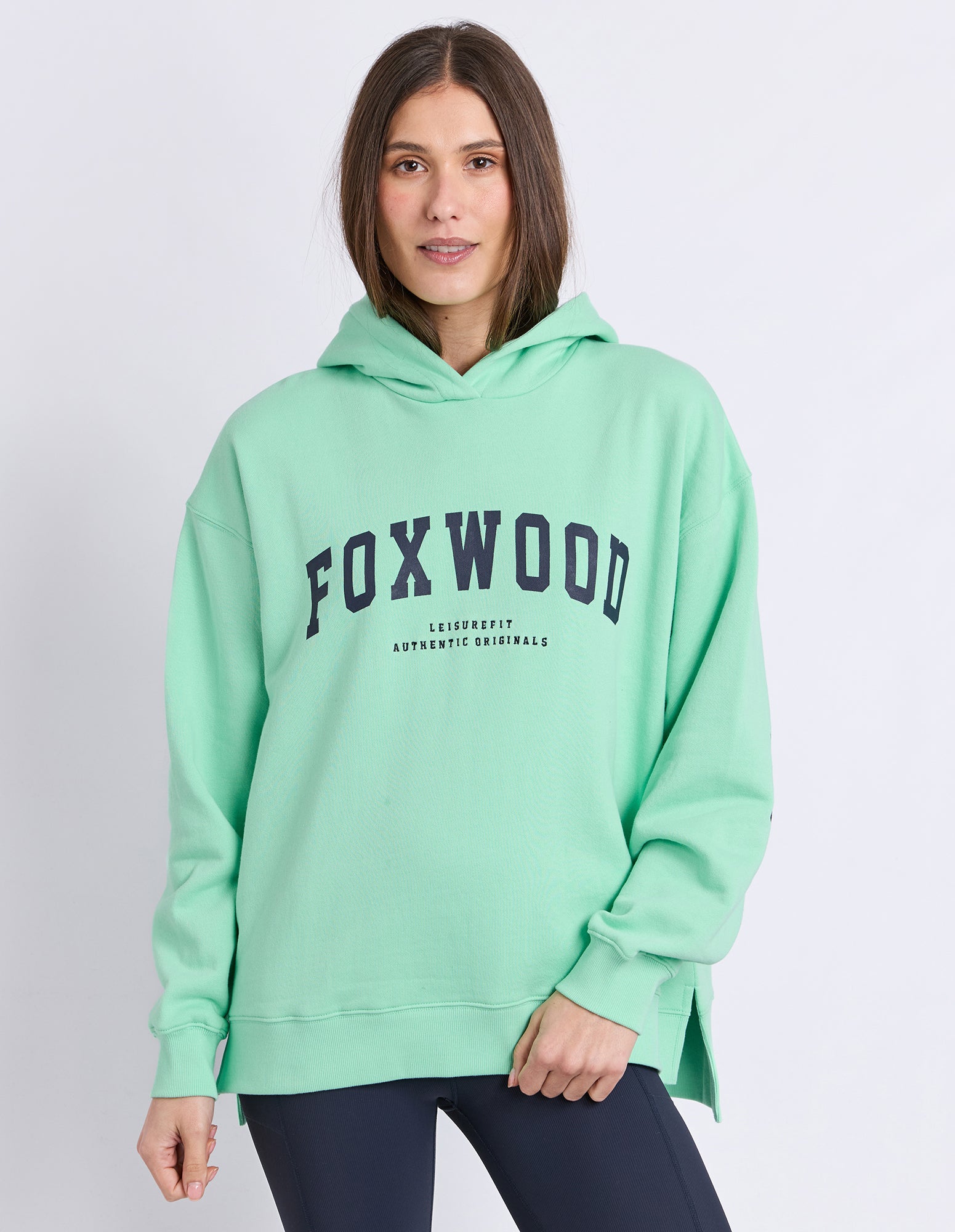 A woman wearing a neon mint green hooded jumper with 'FOXWOOD' printed in navy blue across the chest, paired with dark leggings, against a light grey studio background.