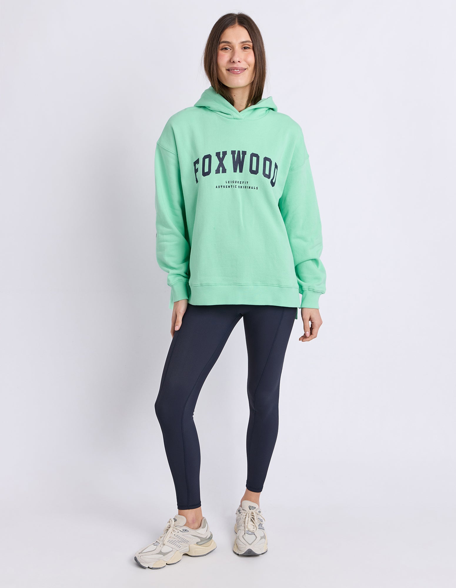 A woman modeling a neon mint green Foxwood hoodie with dark navy lettering on the chest, paired with dark navy leggings and beige sneakers, against a light grey studio background.
