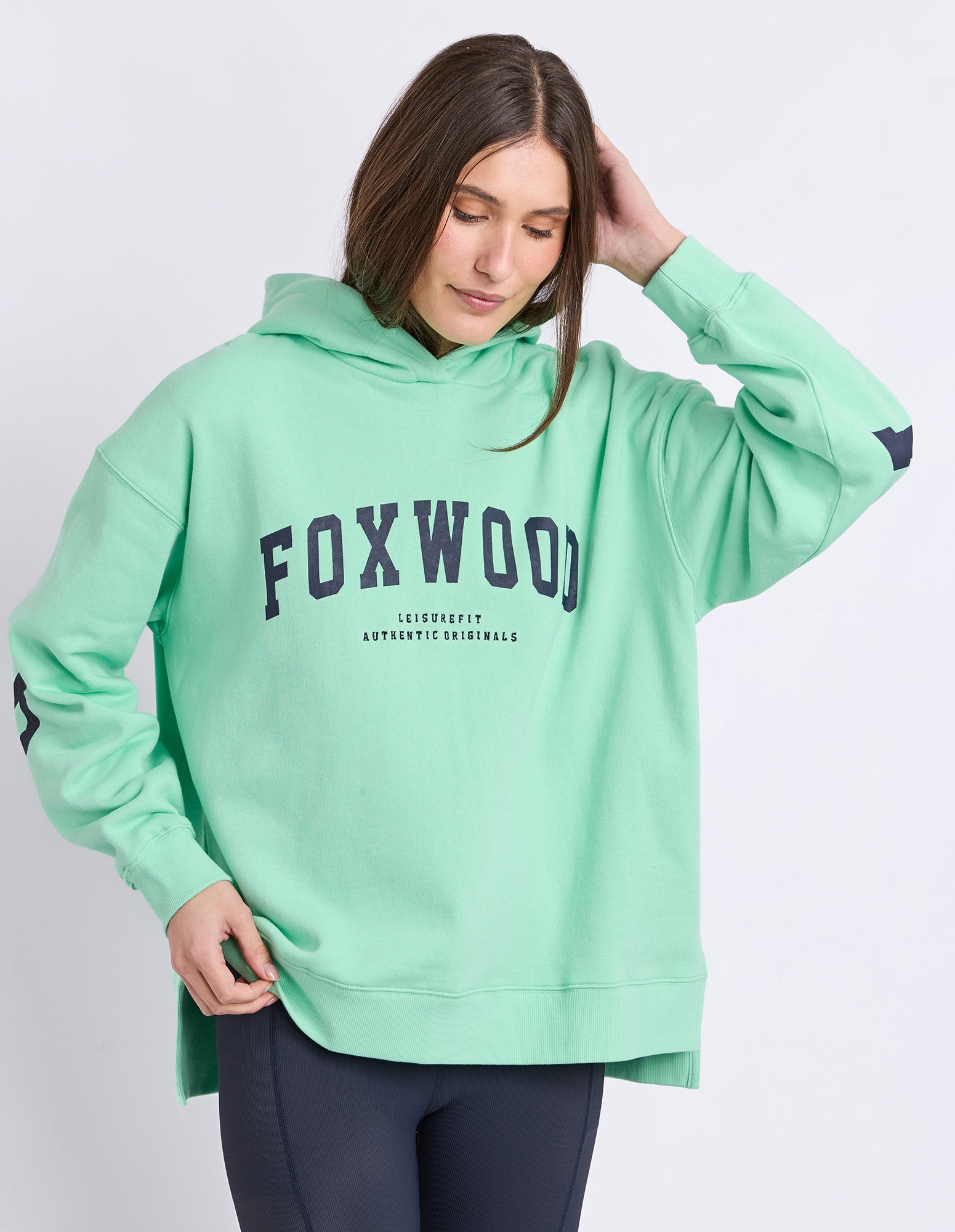 A woman wearing a neon mint green hooded jumper with 'FOXWOOD' printed in navy blue across the chest, paired with dark leggings, against a light grey studio background.