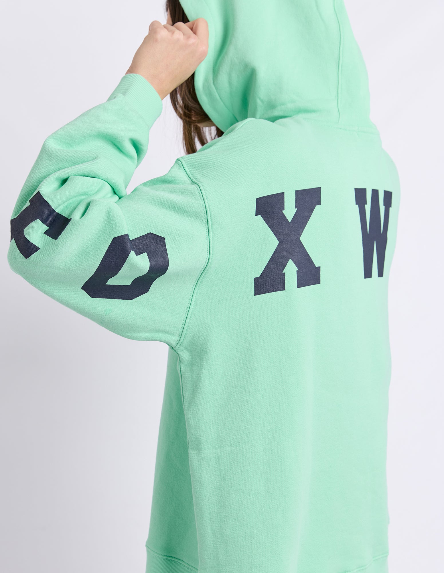 A neon mint green hoodie with large dark navy 'FOXWOOD' printed across the back, shown on a model from a rear three-quarter view against a light grey studio background.