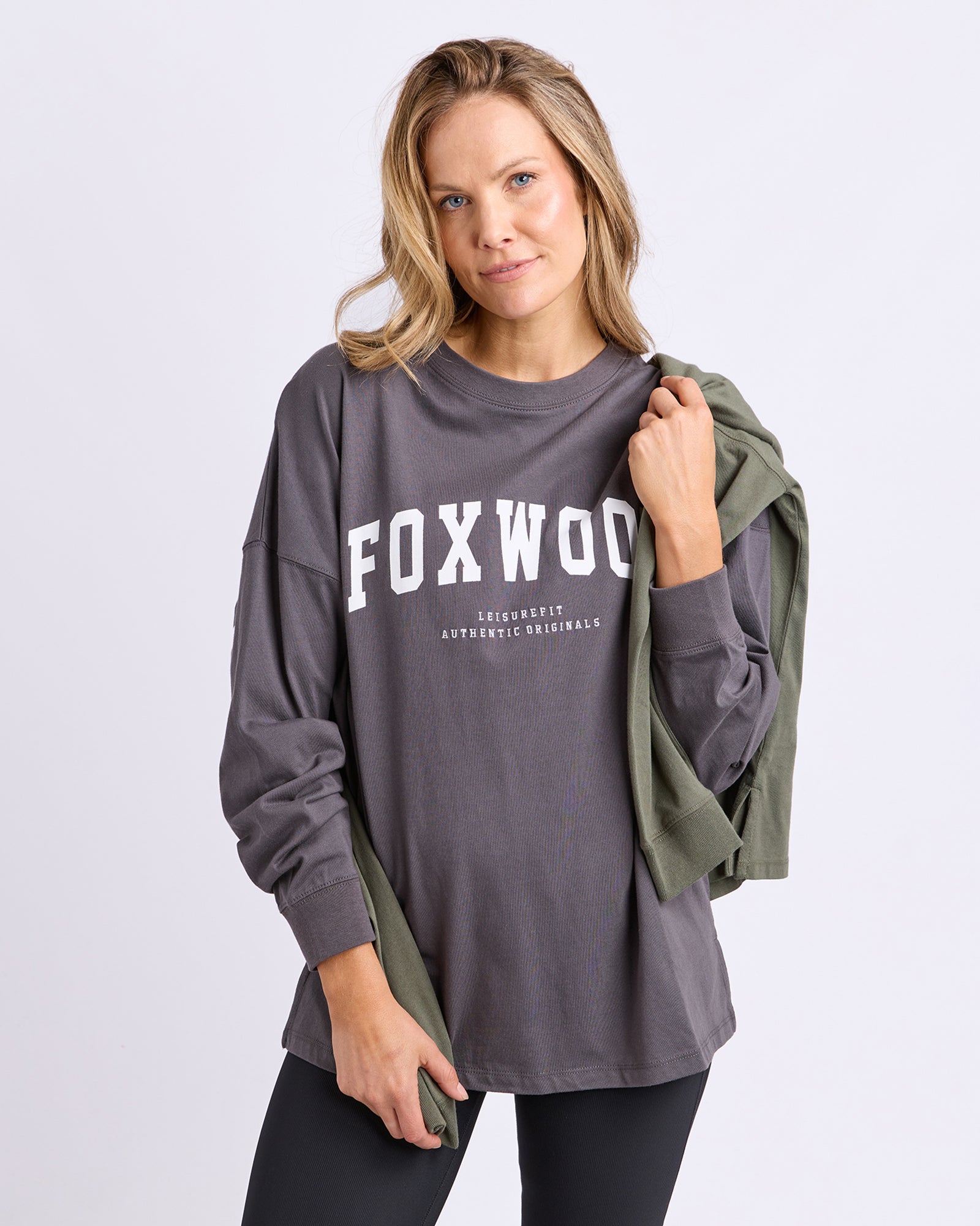 A woman modeling a dark gray long-sleeve t-shirt with 'FOXWOOD' printed in white across the chest, while holding an olive green shirt over her shoulder against a light grey studio background.