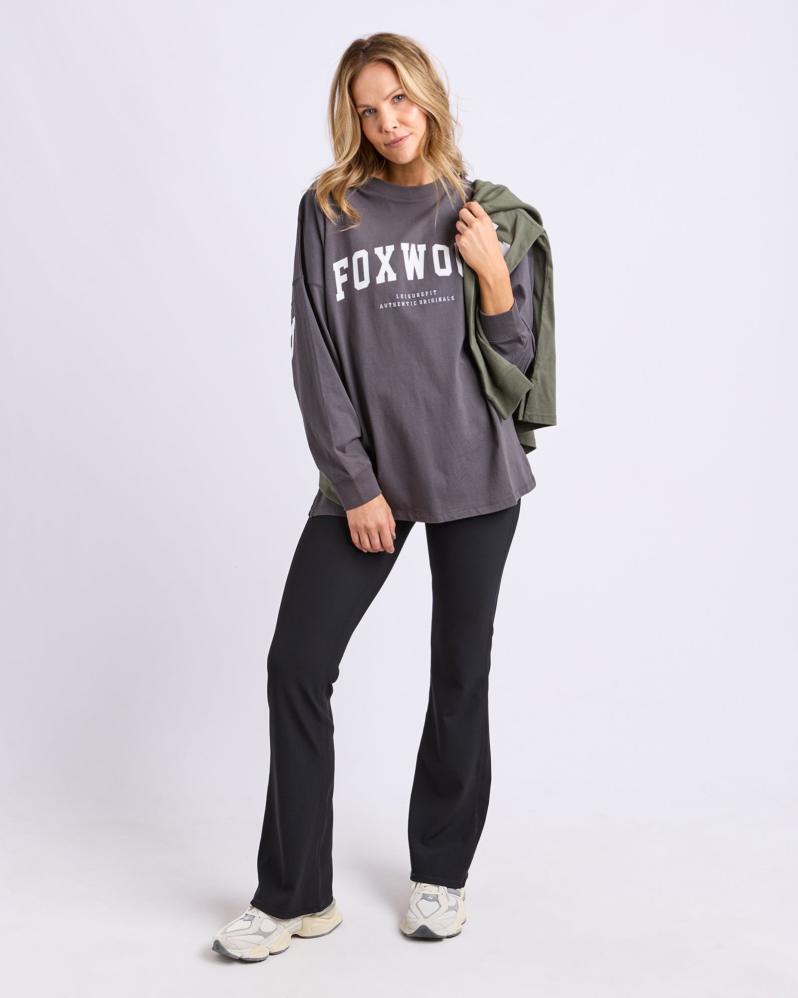 Image of A woman modeling a dark grey oversized long-sleeve tee with 'FOXWOOD' printed in white across the chest, paired with black pants and white sneakers, while holding an olive green jacket over her shoulder against a light grey studio background.