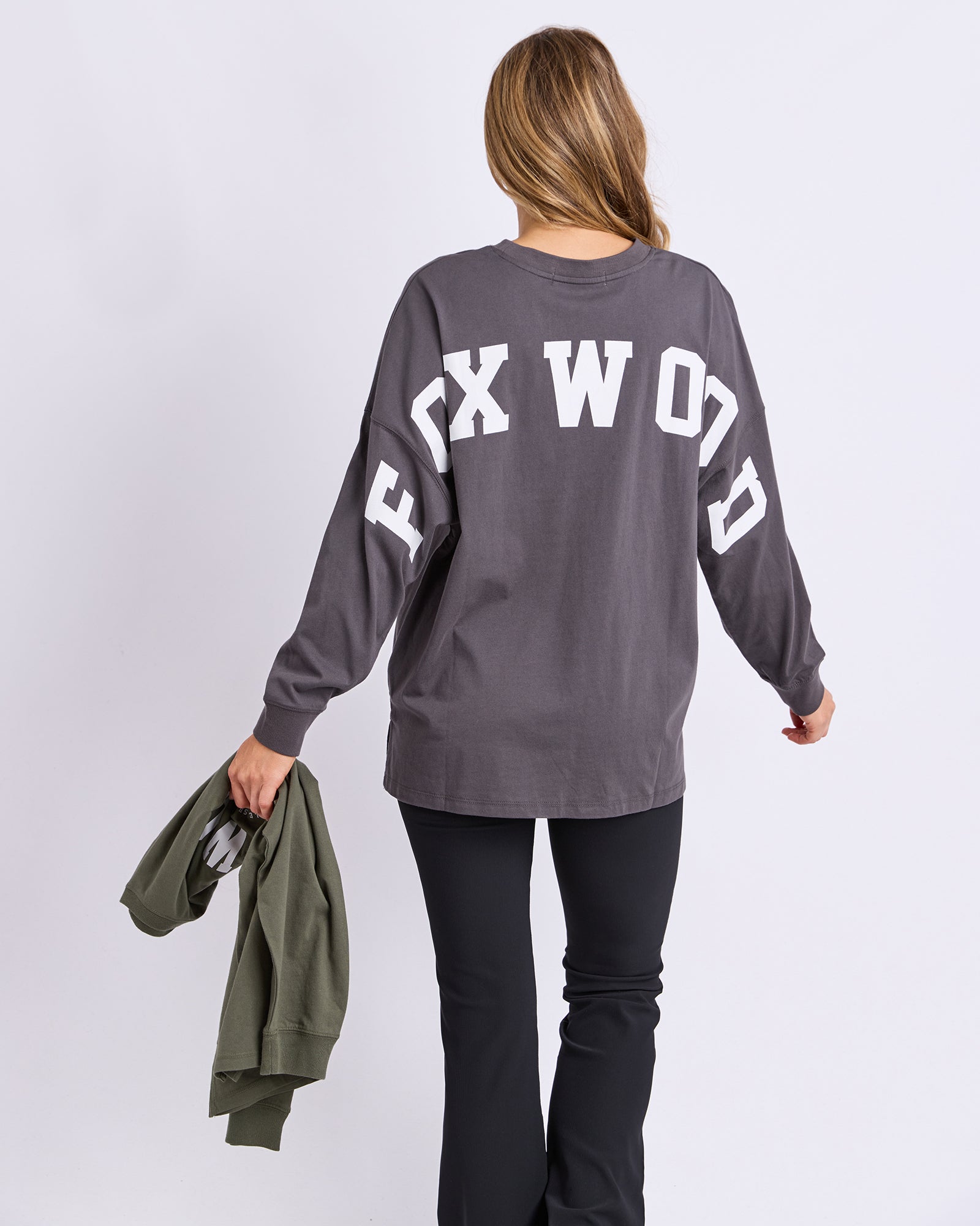 A woman shown from the back wearing a dark gray long-sleeve tee with 'FOX WOOD' printed in large white letters across the back, paired with black pants and holding an olive green sweatshirt against a light grey studio background.