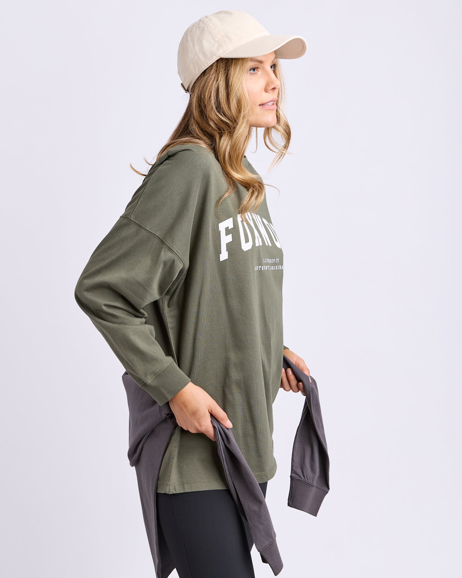 Image of Woman wearing a khaki long sleeve tee with Foxwood in white, holding a grey tee against a white background