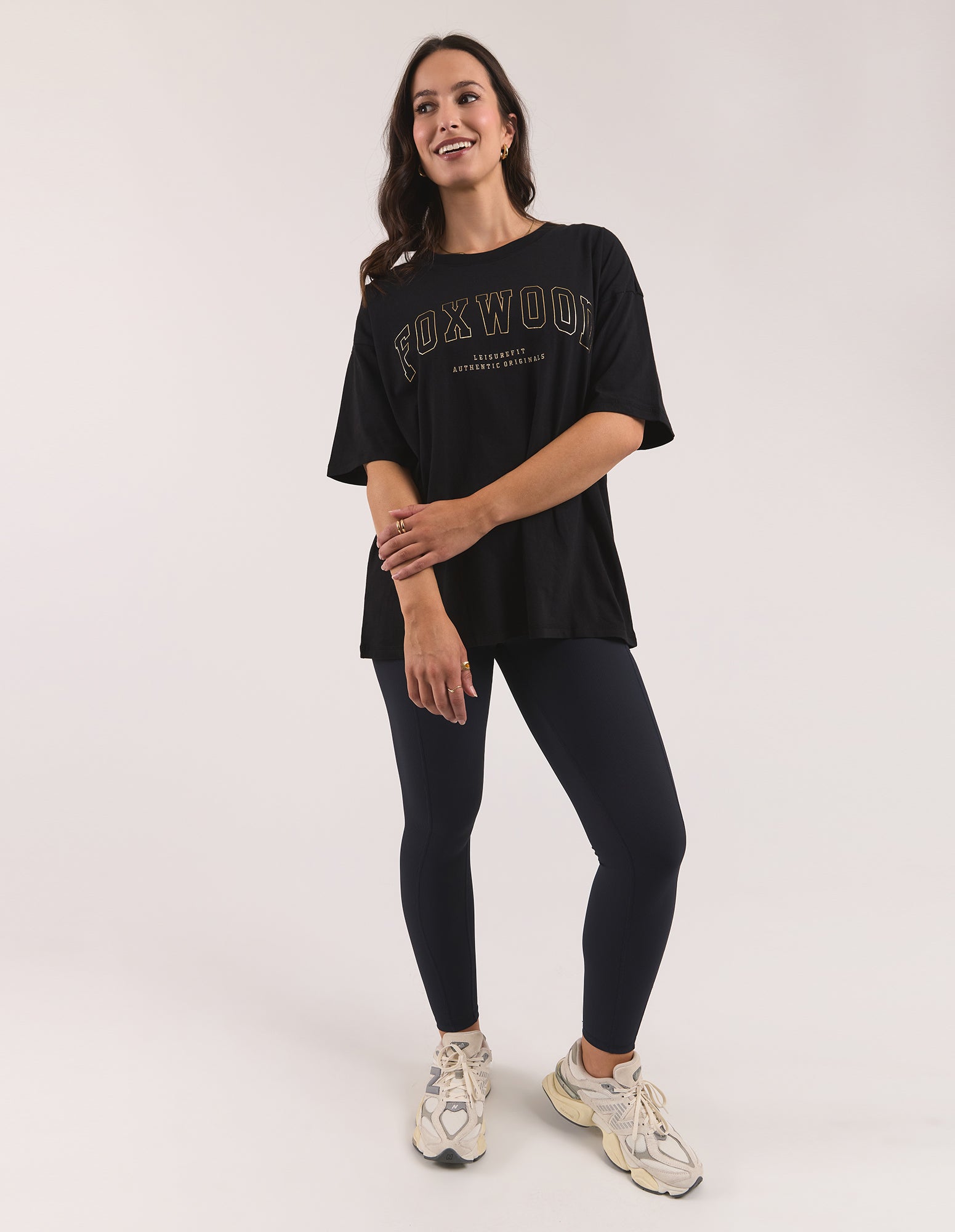 Image of A woman modeling a black Foxwood short-sleeve graphic t-shirt paired with black leggings and beige sneakers, standing against a plain light grey studio background.