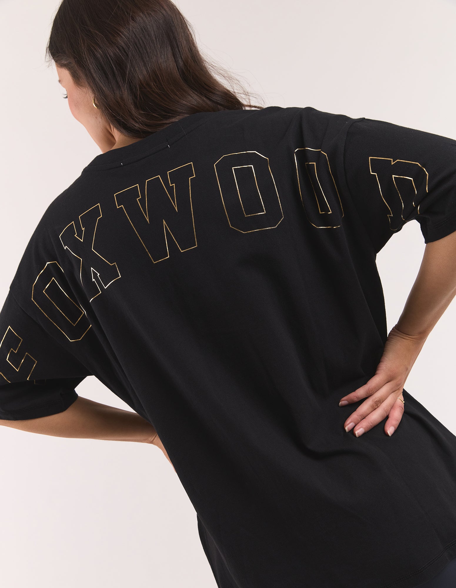Image of A woman wearing an oversized black T-shirt with 'FOXWOOD' printed across the back in a gold outline font, shown from a rear three-quarter angle against a plain light background.