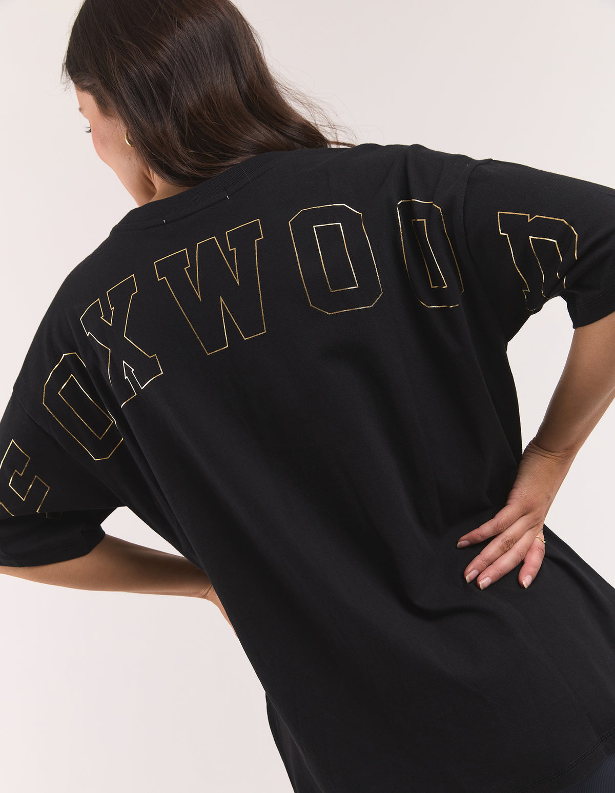 A woman wearing an oversized black T-shirt with 'FOXWOOD' printed across the back in a gold outline font, shown from a rear three-quarter angle against a plain light background.