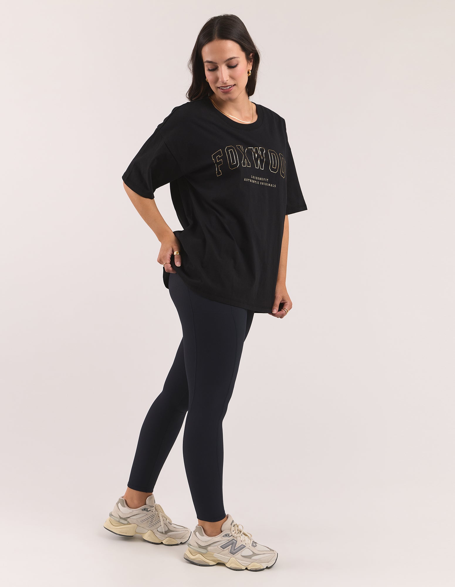 Image of A woman modeling an oversized black t-shirt with 'FOXWOOD' printed in gold outline across the chest, paired with dark leggings and New Balance sneakers, against a light grey studio background.