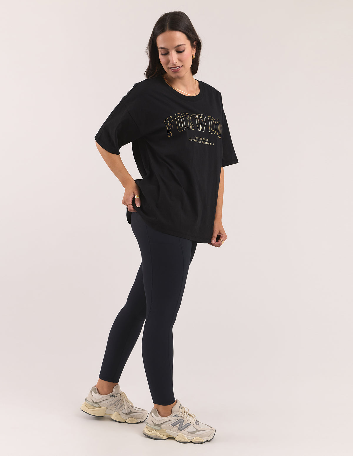 A woman modeling an oversized black t-shirt with 'FOXWOOD' printed in gold outline across the chest, paired with dark leggings and New Balance sneakers, against a light grey studio background.
