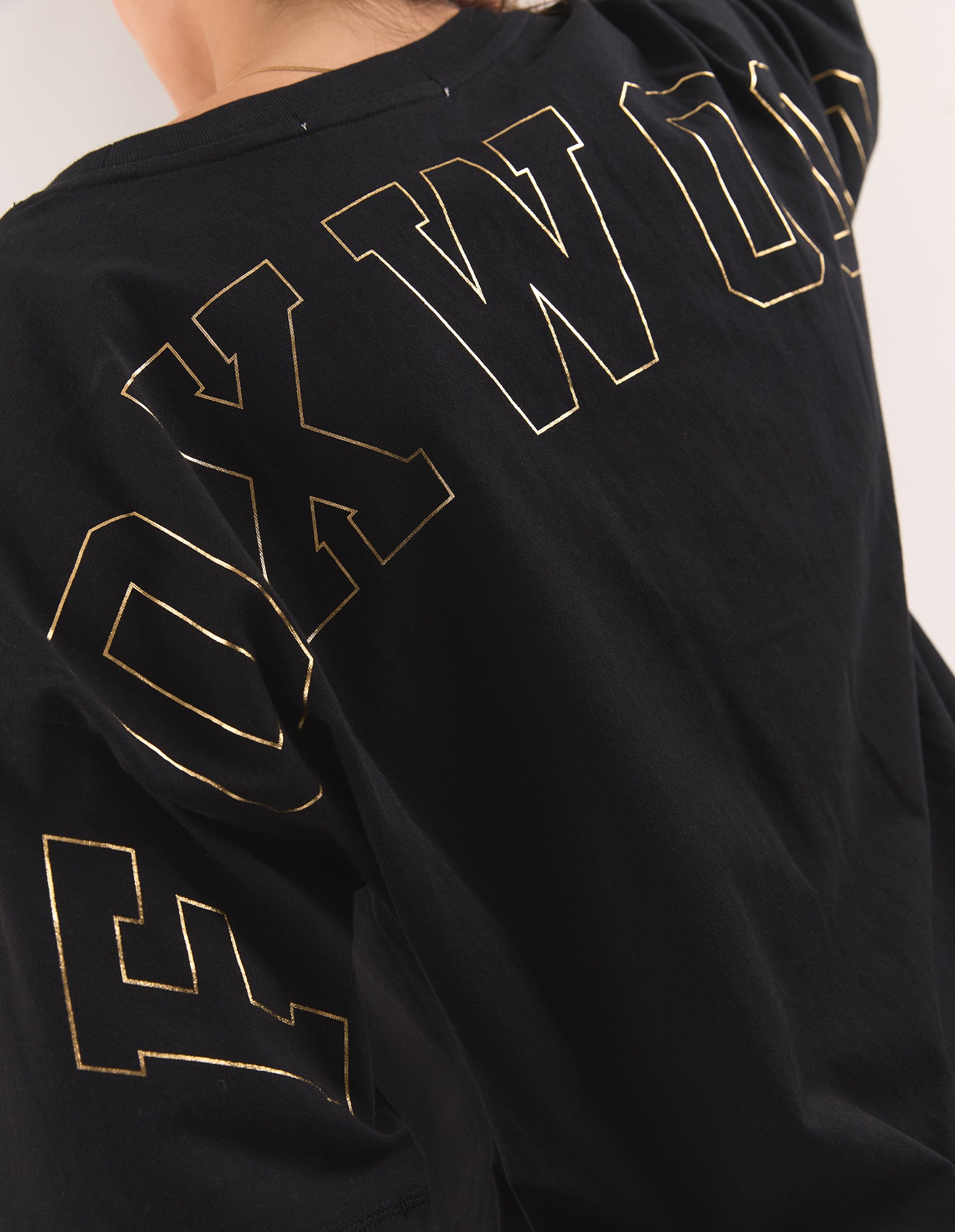 Image of Close-up of a woman wearing a black oversized T-shirt with large gold outlined varsity-style lettering across the back.