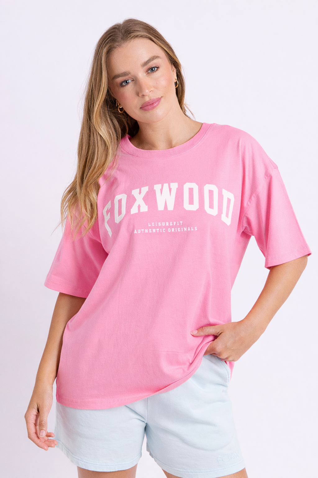 Image of A woman wearing a pink short-sleeve T-shirt with 'FOXWOOD' printed in white across the chest, paired with light blue shorts, against a plain light background.