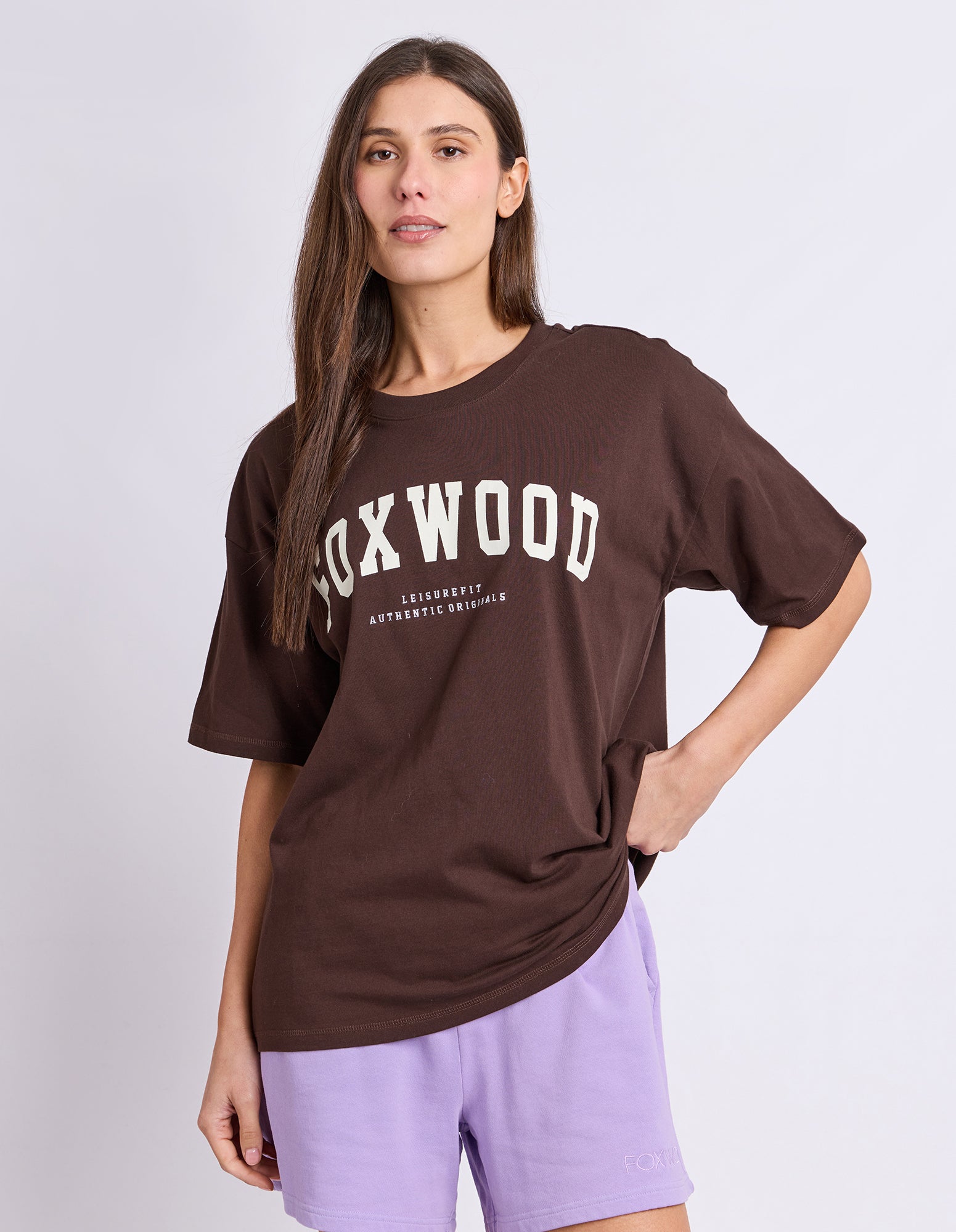 Image of A woman wearing an oversized brown t-shirt with 'FOXWOOD' printed in white across the chest, paired with lavender shorts, against a light grey studio background.