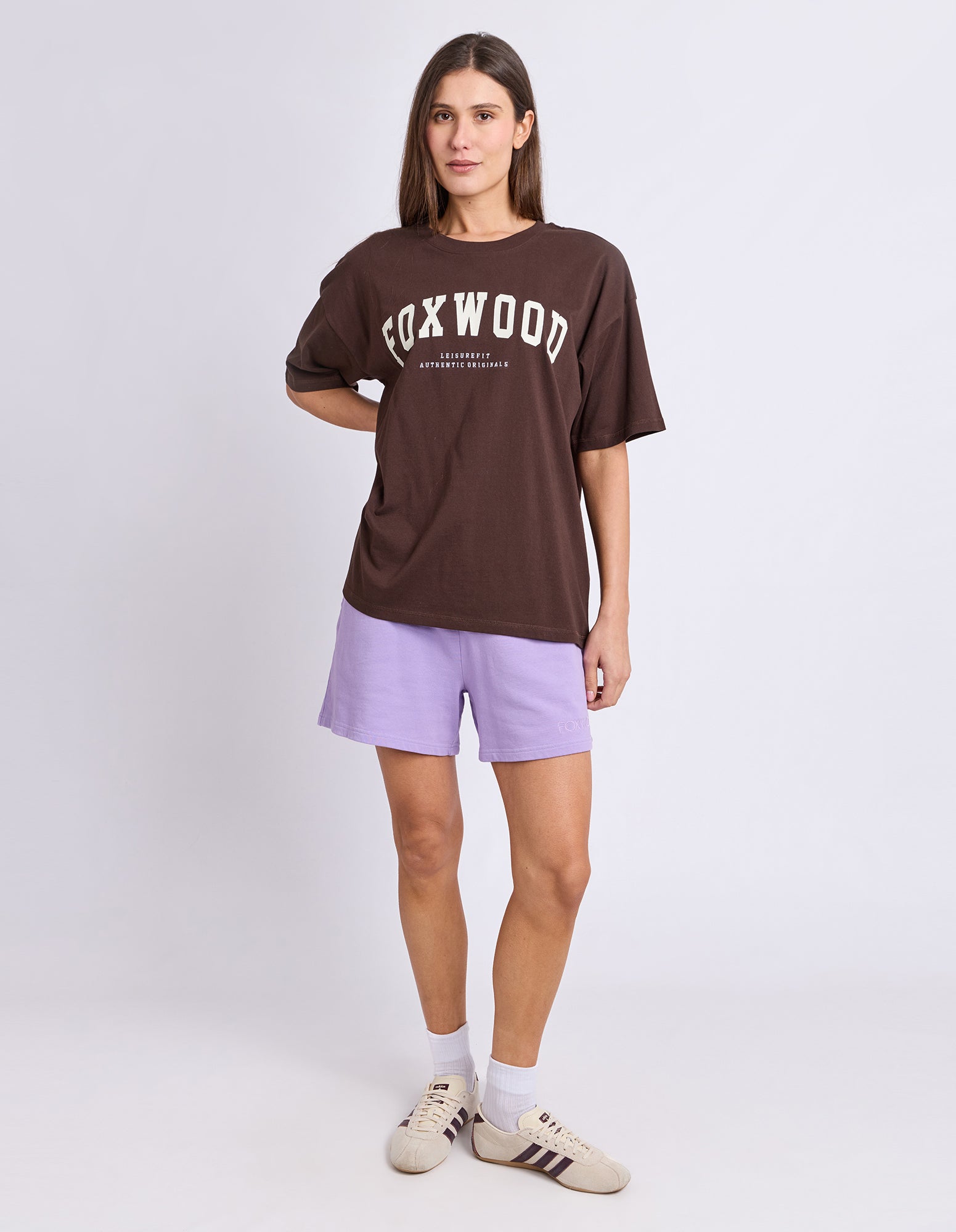 A woman wearing an oversized brown t-shirt with 'FOXWOOD' printed in white across the chest, paired with light purple shorts, white socks, and beige sneakers, against a light grey studio background.