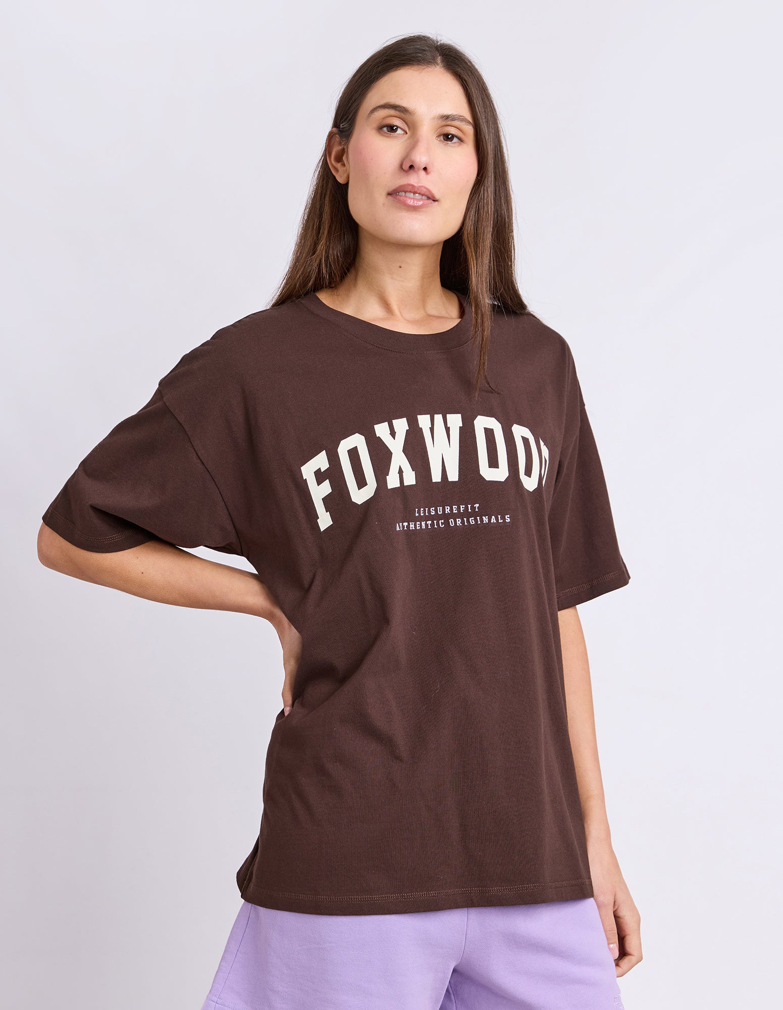 Image of A woman wearing an oversized brown t-shirt with 'FOXWOOD' printed in white across the chest, paired with light purple sweatpants against a light grey studio background.