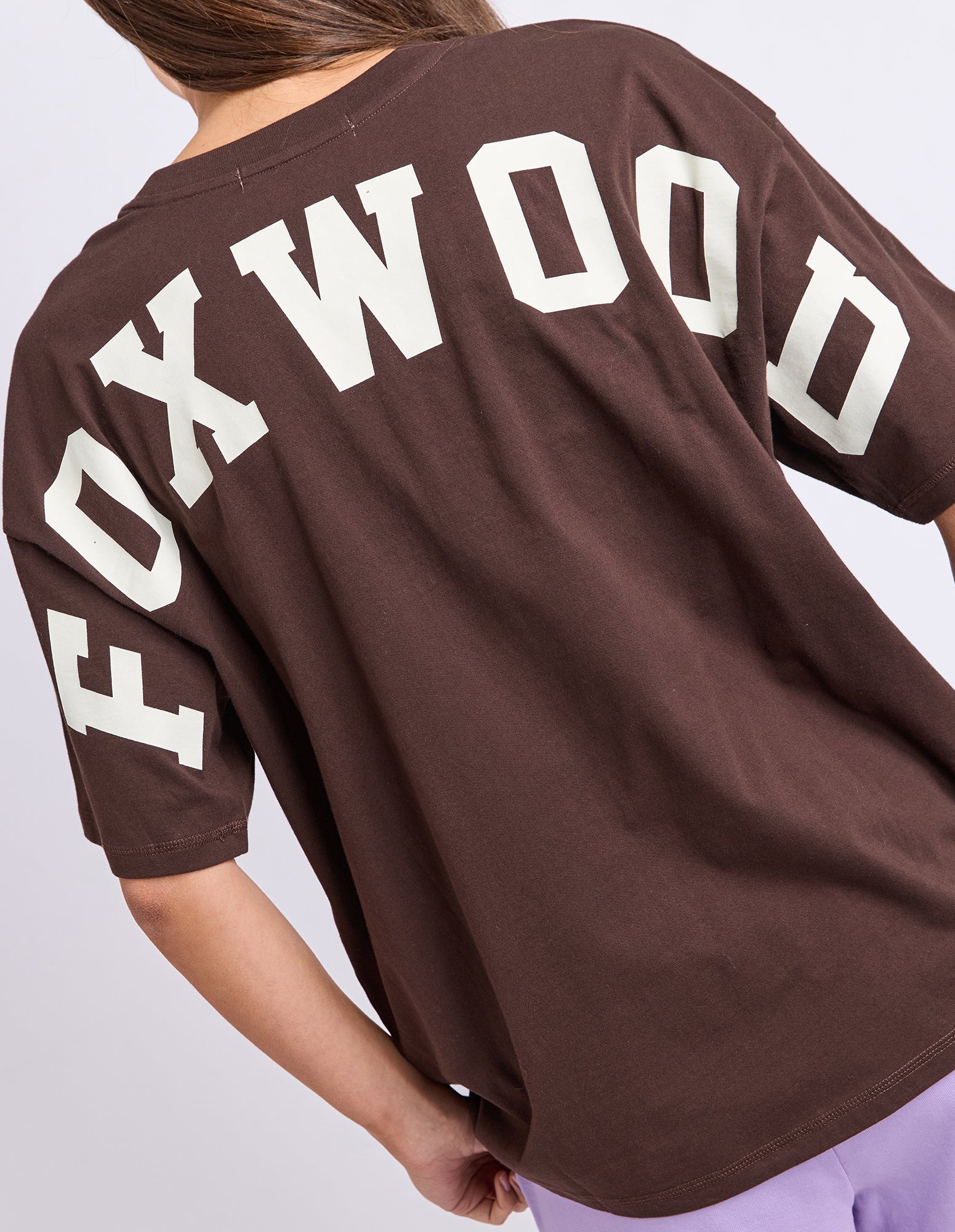 Image of A close-up back view of a woman wearing an oversized brown T-shirt with large white 'FOXWOOD' lettering across the upper back and sleeves, paired with light purple bottoms.