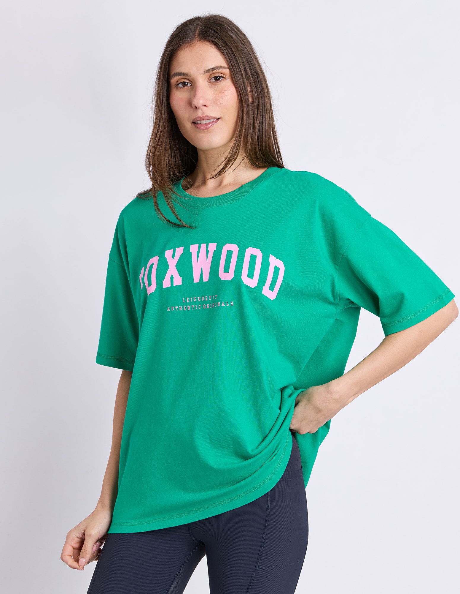 Image of A woman wearing an oversized green t-shirt with 'FOXWOOD' printed in pink on the chest, paired with dark leggings, against a light grey studio background.