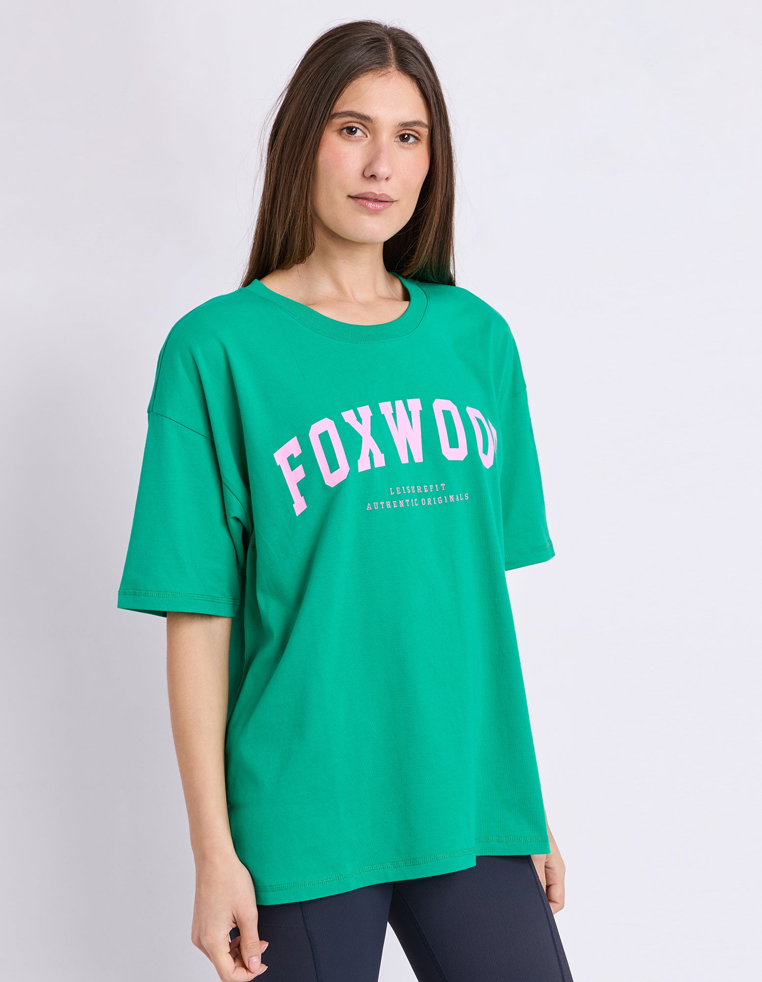 A woman wearing an oversized green t-shirt with 'FOXWOOD' printed in pink, set against a plain light grey background.
