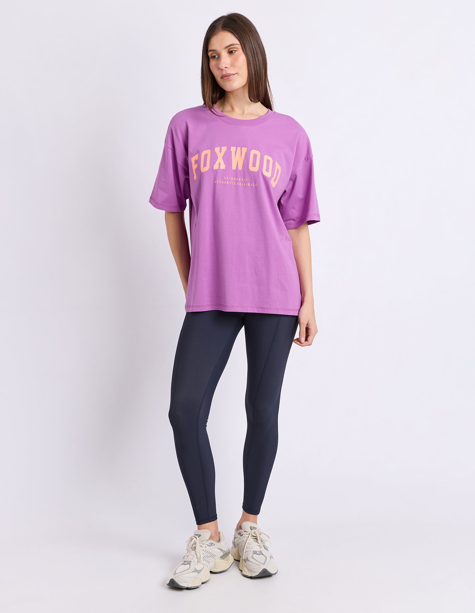 Image of A woman modeling an oversized purple Foxwood graphic t-shirt paired with dark leggings and white sneakers, standing against a plain white studio background.