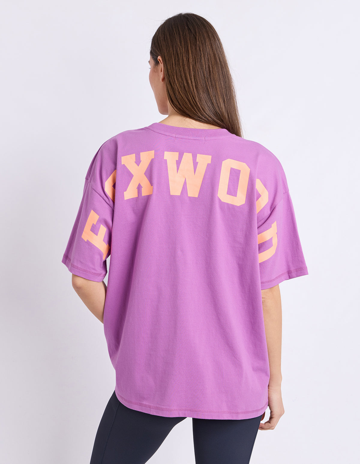 A woman viewed from the back wearing an oversized purple t-shirt with 'FOXWOOD' printed in orange across the back, paired with dark leggings, against a plain white studio background.
