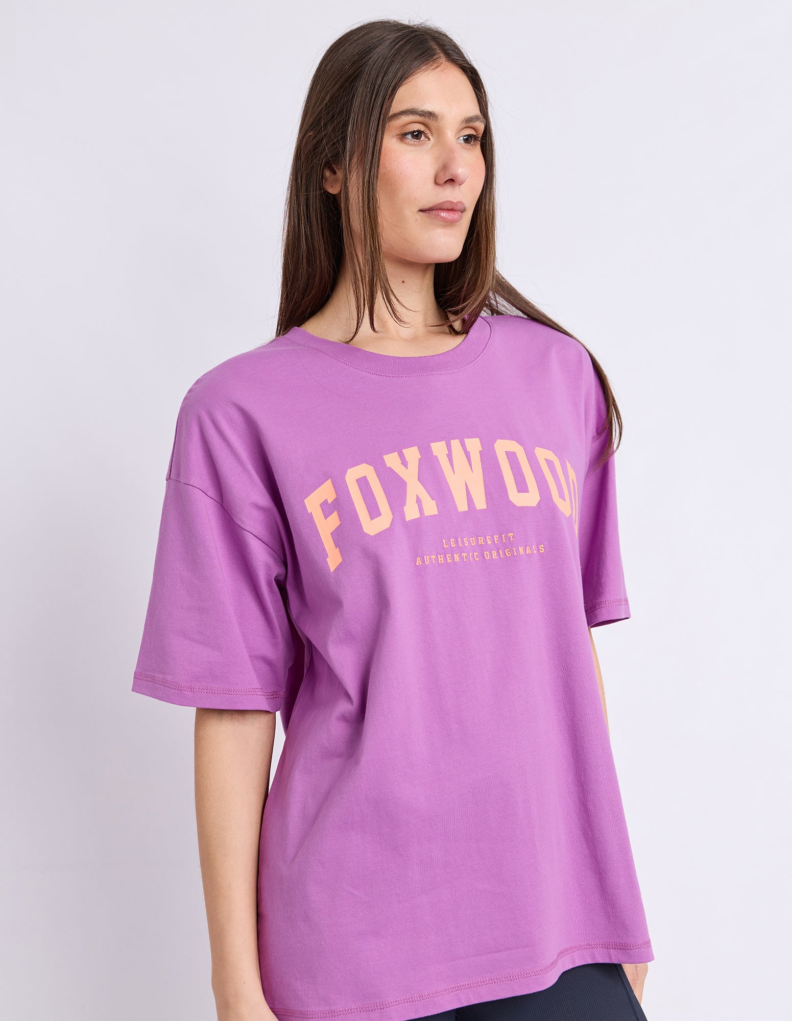 Image of A woman wearing a purple Foxwood t-shirt with an orange graphic text print on the front, set against a plain light grey studio background.