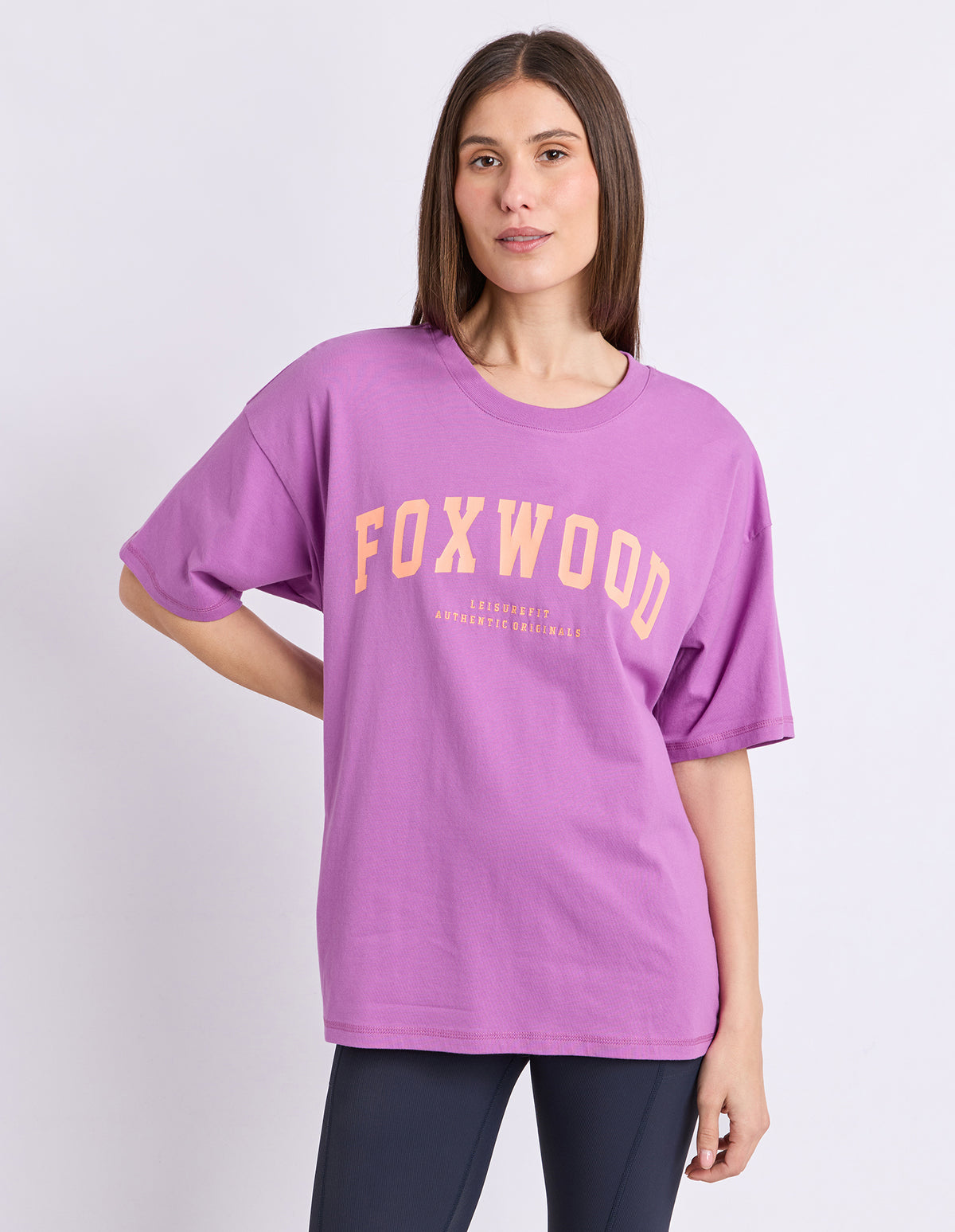 A woman modeling an oversized purple t-shirt with 'FOXWOOD' printed in orange arched font across the chest, paired with dark leggings, against a light grey studio background.
