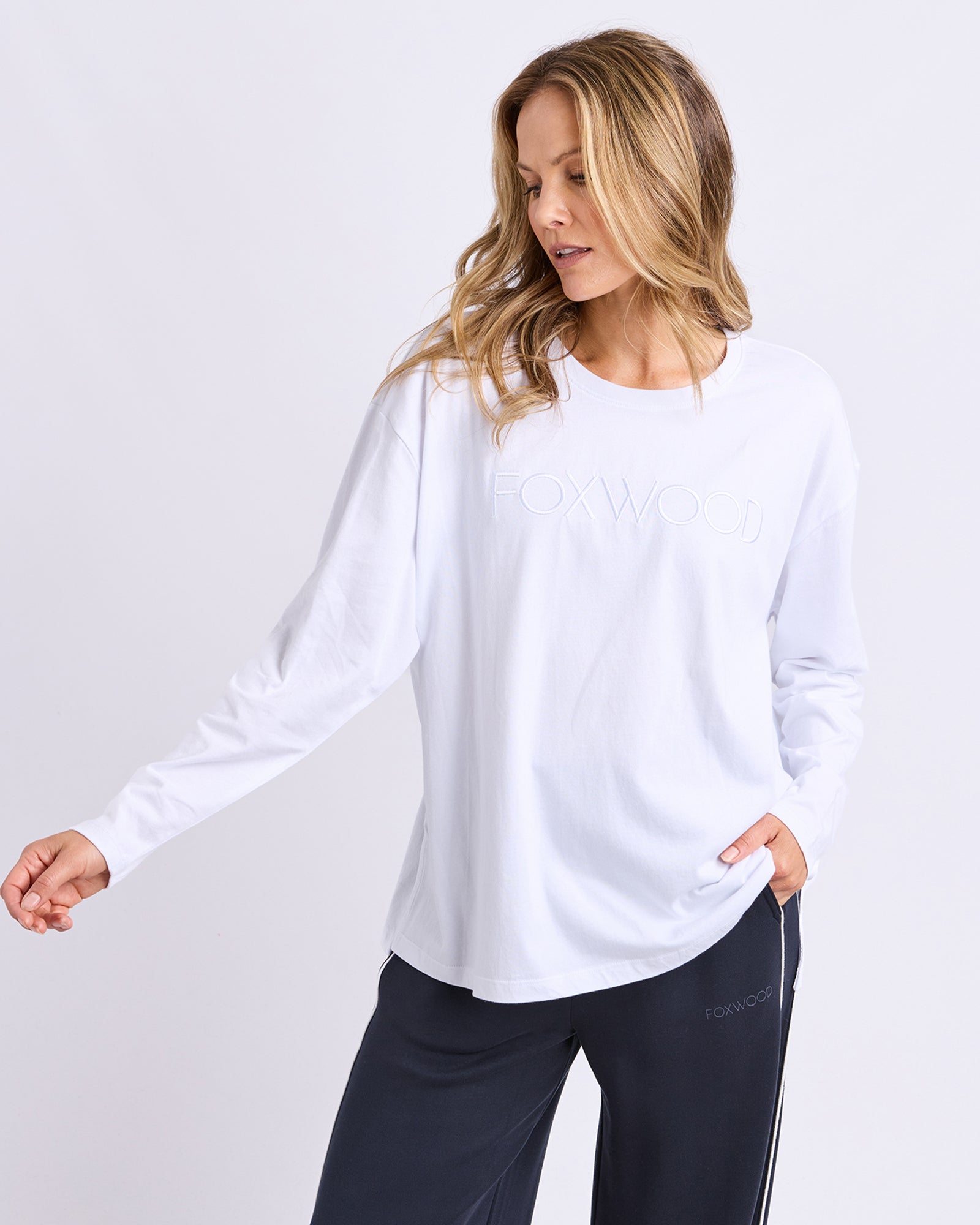 Woman wearing a white long-sleeve top with Foxwood embroidered on the front and black pants on a light grey background