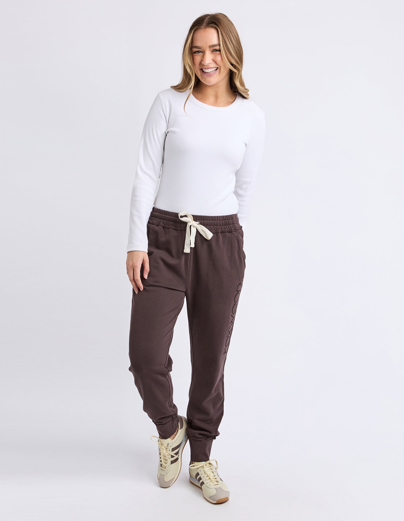 Image of Woman wearing a white long-sleeve top and brown track pants on a white background