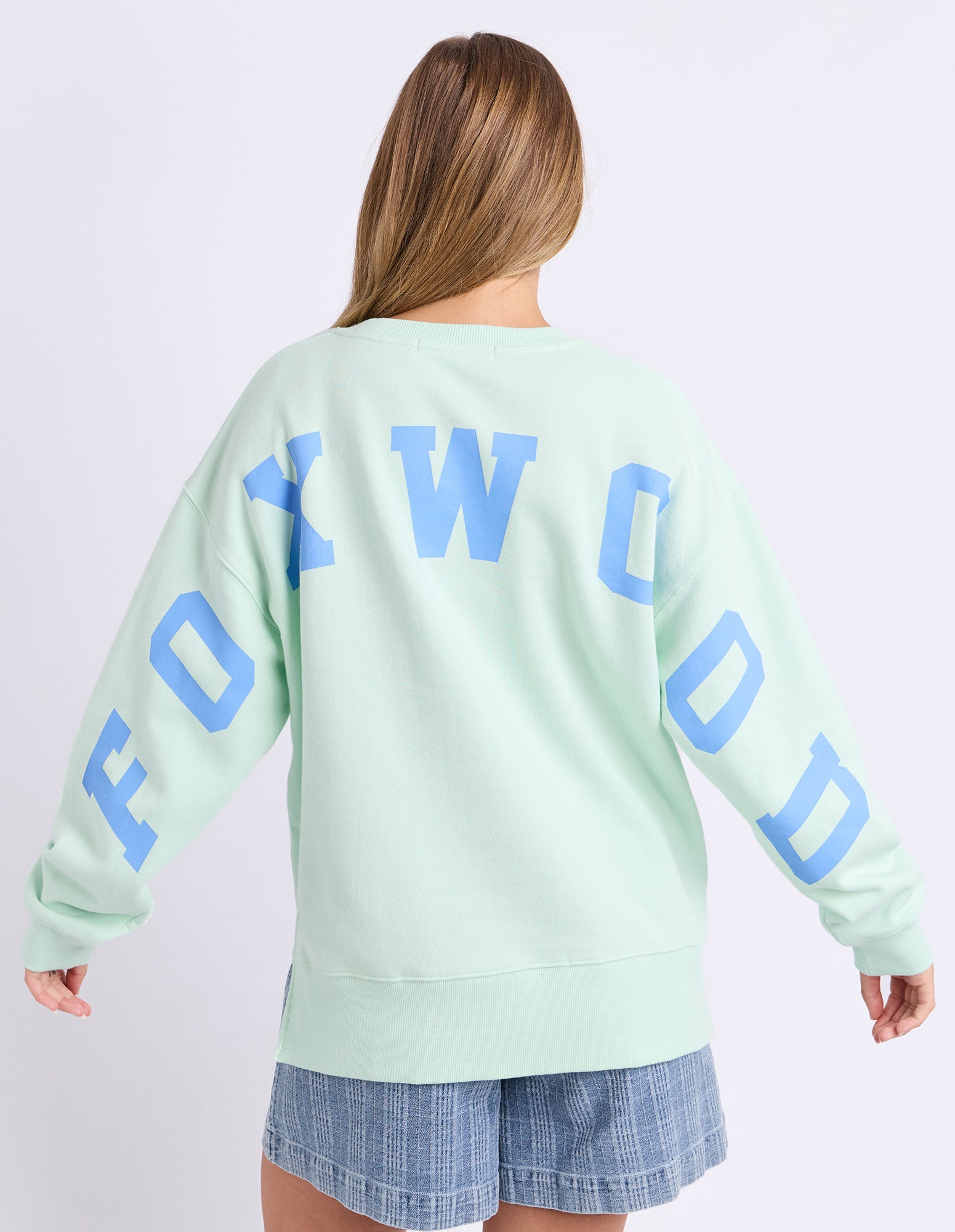 Image of Woman wearing a mint green jumper with 'FOXWOOD' printed on the back in blue against a white background