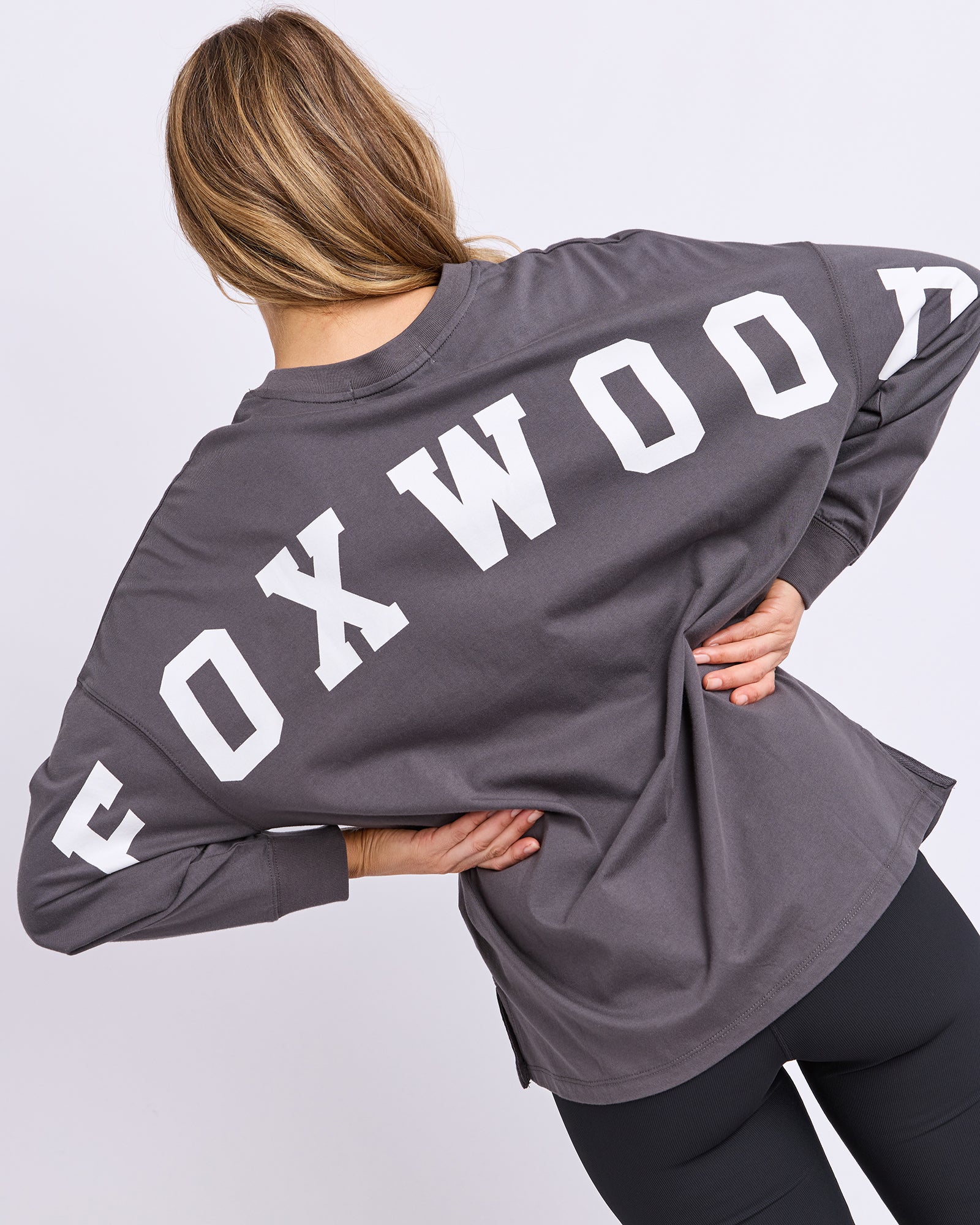 Image of A woman shown from the back wearing a dark grey long-sleeve tee with 'FOXWOOD' printed in large white letters across the back, paired with black leggings, against a light grey studio background.