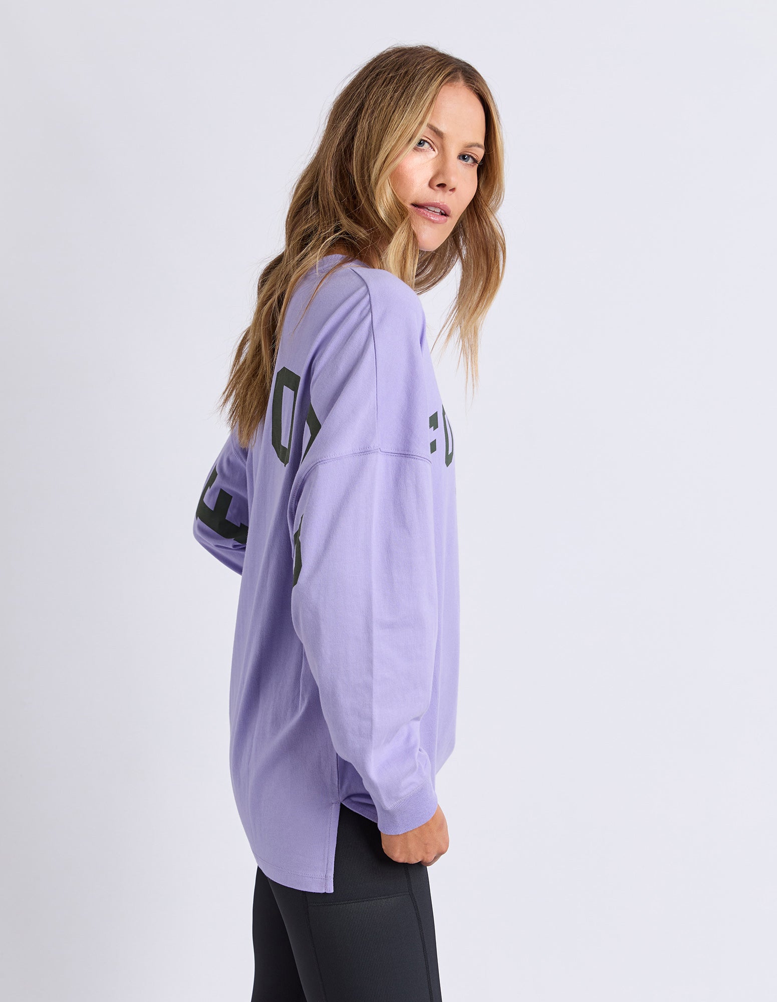 Sideview of a woman wearing a light purple oversized long sleev tee with khaki text on a white background