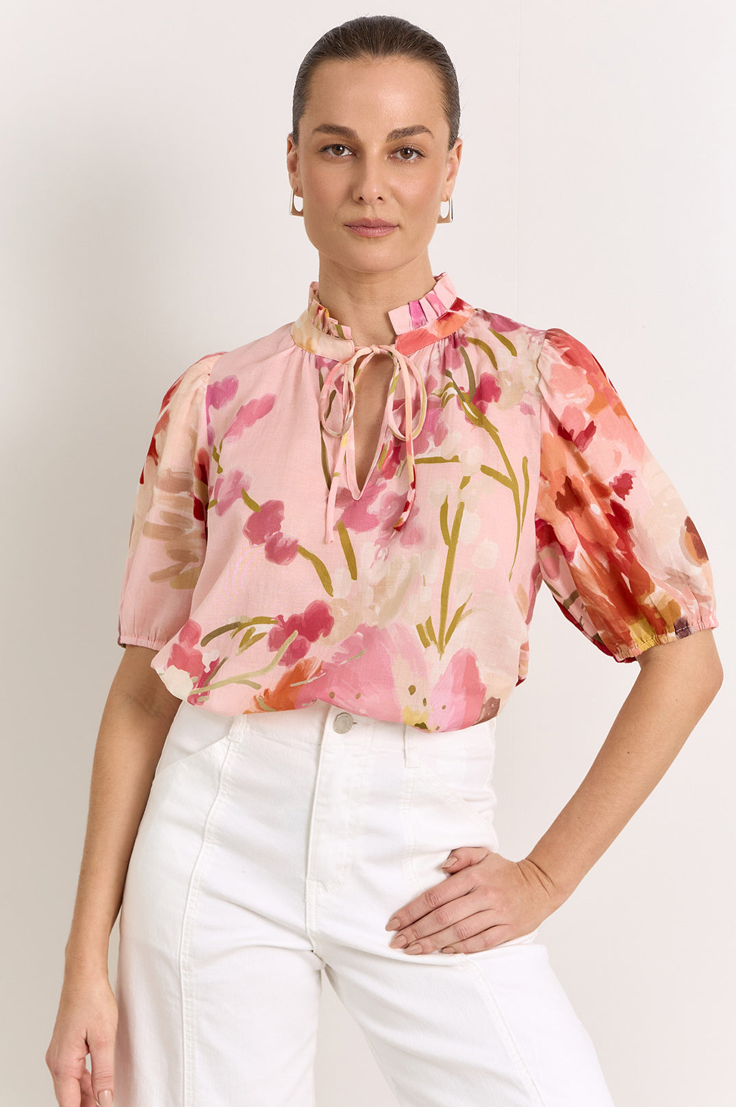 A woman wearing a light pink short-sleeve blouse with a water colour floral print in shades of red, orange, and green, paired with white trousers, against a light grey studio background.